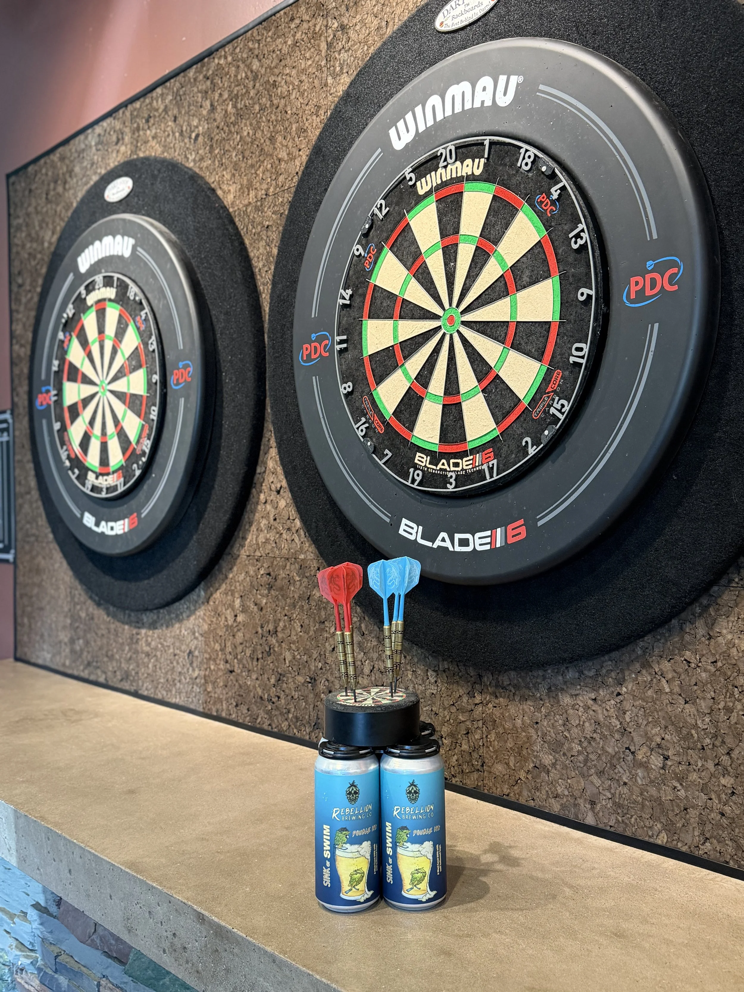 Two dartboards mounted on a cork wall, with two darts each in the bullseye, and two sink or swim DIPA beer cans of Rebellion Brewing Co. beverage cans on a platform below.
