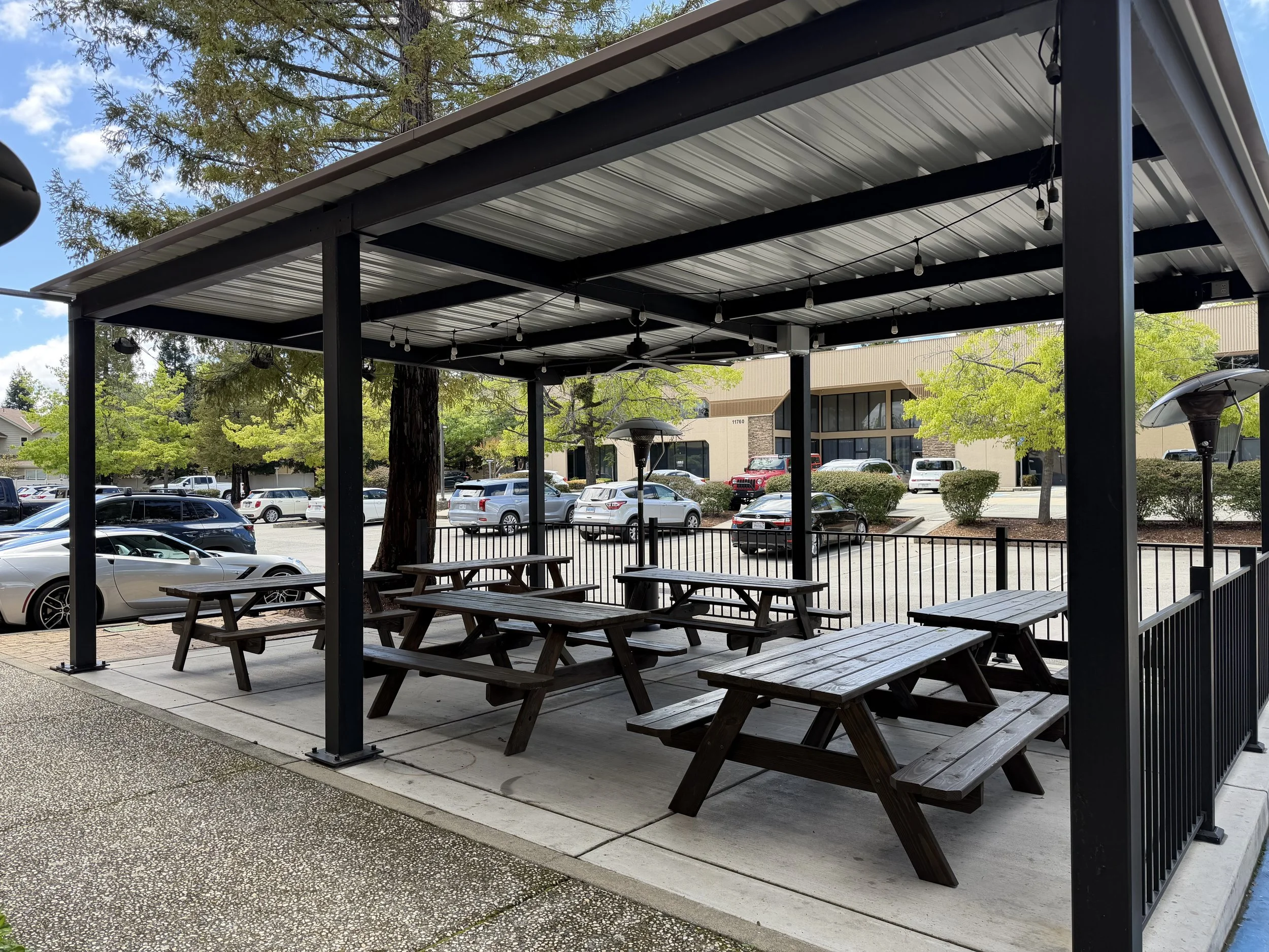 Outdoor patio seating area with wooden picnic tables and a metal roof, surrounded by a parking lot with cars and trees.