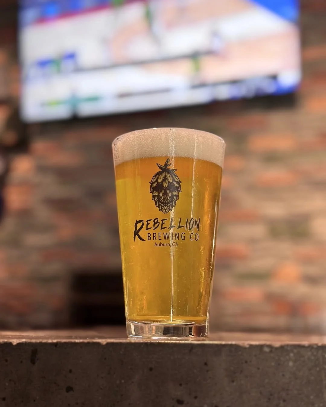 A pint glass of beer with foam head and logo of Rebel Lion Brewing Co., Auburn, CA, on a wooden surface in front of a blurred brick wall and TV screen.