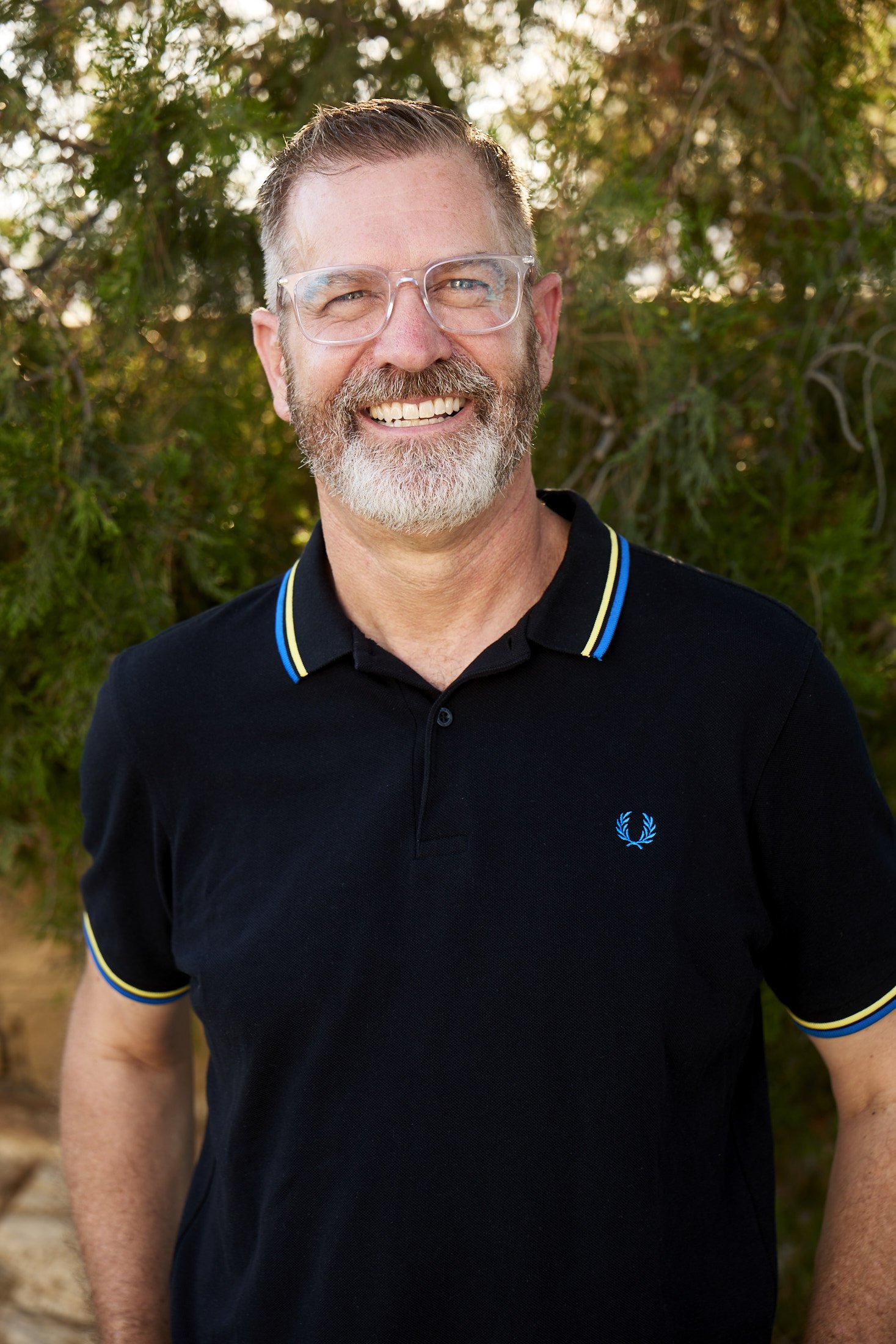 Photographer Andy McRory is smiling, with glasses, a beard, and short hair, wearing a black polo shirt with yellow and blue trim, standing outdoors with trees in the background.