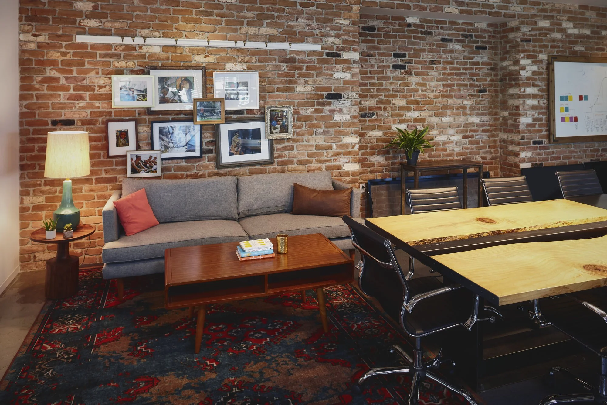 Living room with an exposed brick wall decorated with framed photos, a grey sofa with orange and brown cushions, a wooden coffee table with books and a gold cup, a side table with a lamp and plants, a large rug, and a conference table surrounded by b