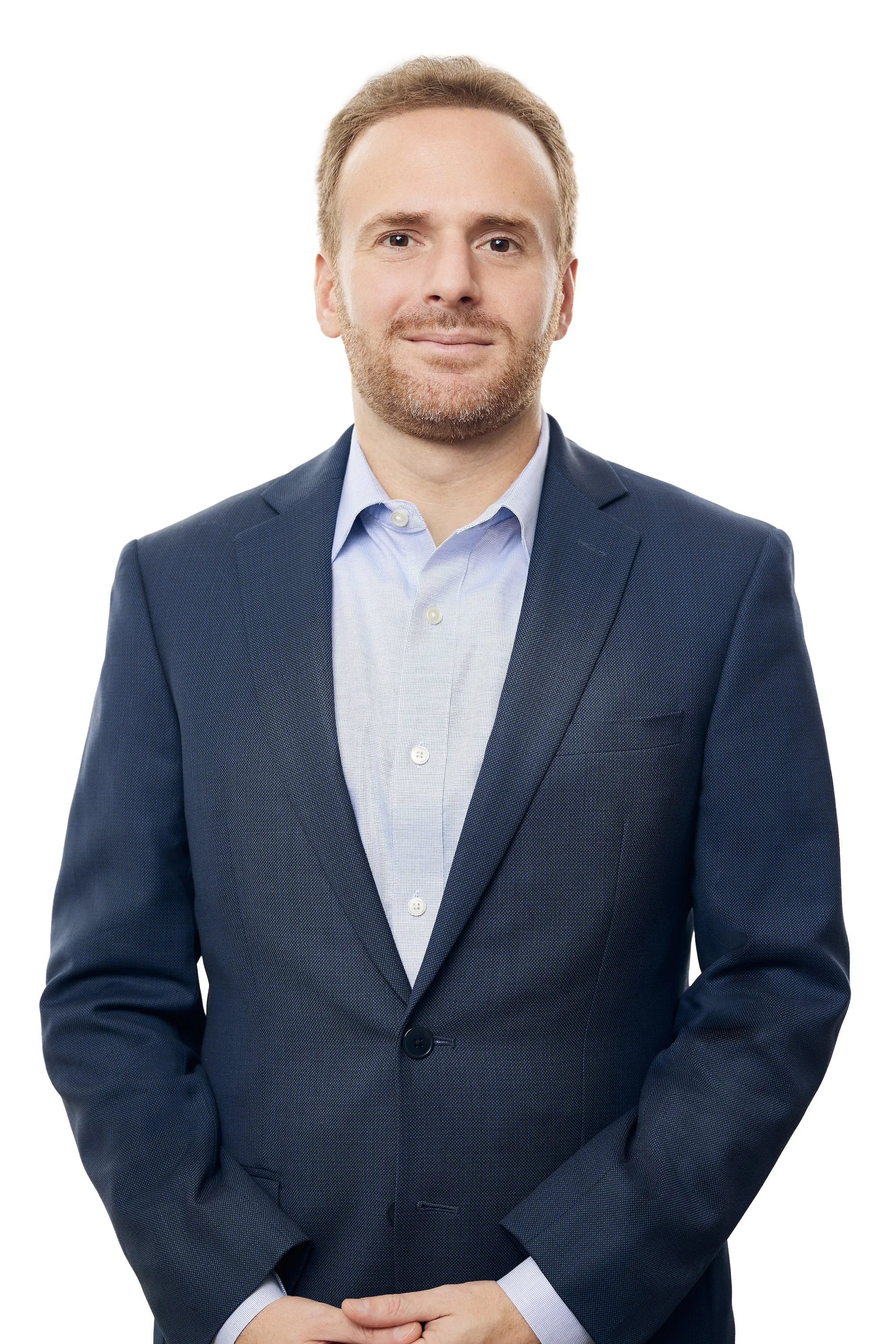 Man with light brown hair and beard in blue shirt and blue jacket, with hands clasped in front