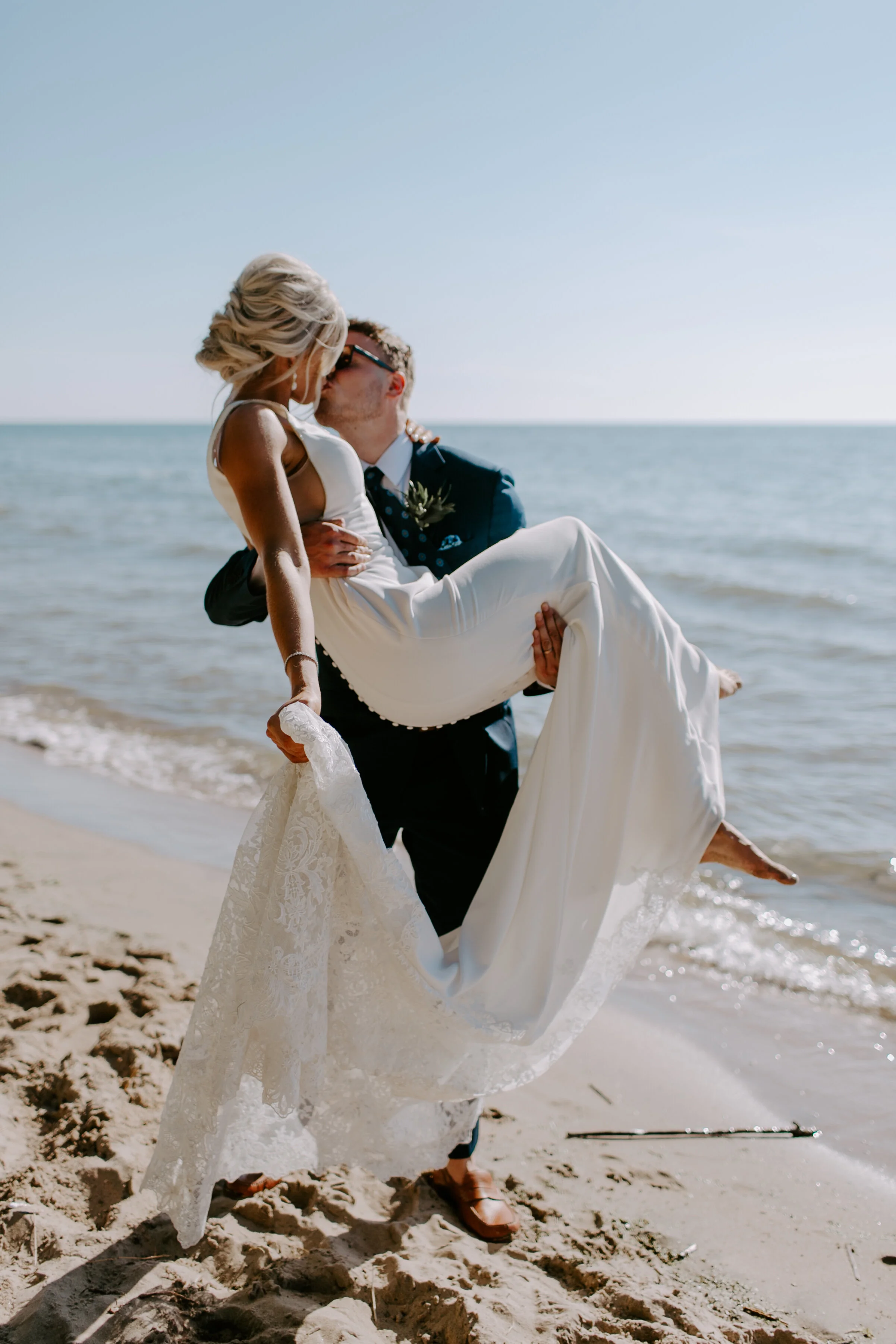 A bride in a white gown being carried by a groom in a suit at the beach, both kissing with the ocean in the background.