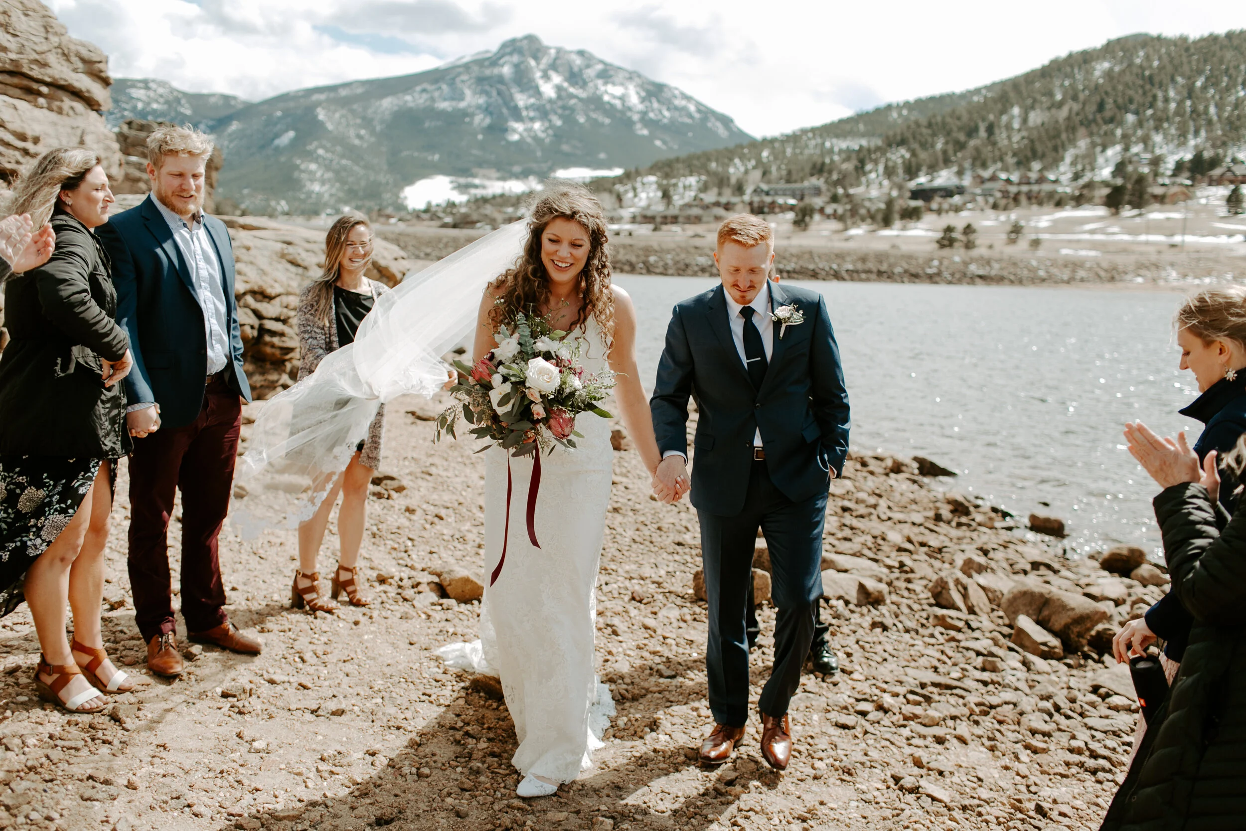 A bride and groom holding hands walking along a rocky lakeside with mountains in the background, surrounded by onlookers clapping and celebrating during a wedding ceremony.