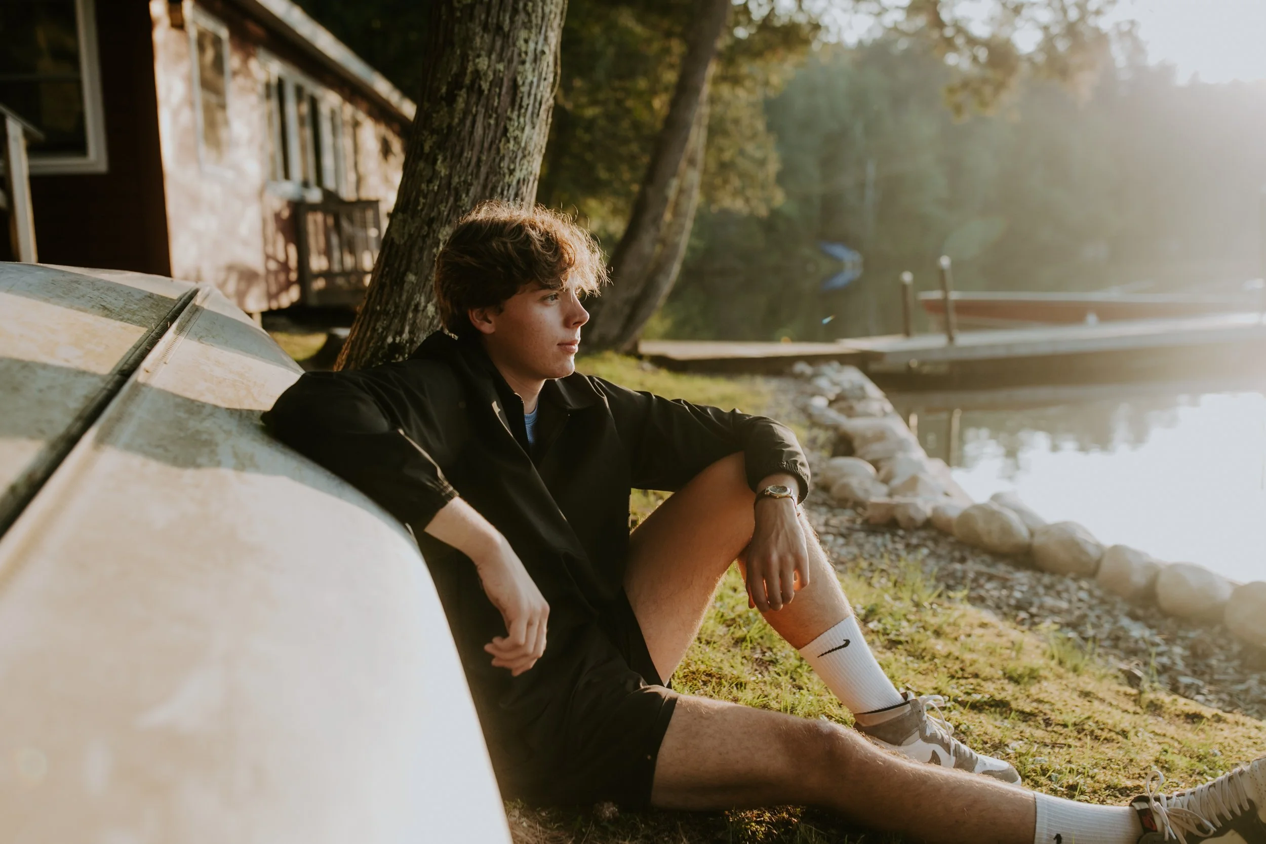 A young man sitting by a lakeshore leaning on an overturned canoe, with trees and a dock in the background, during sunset.