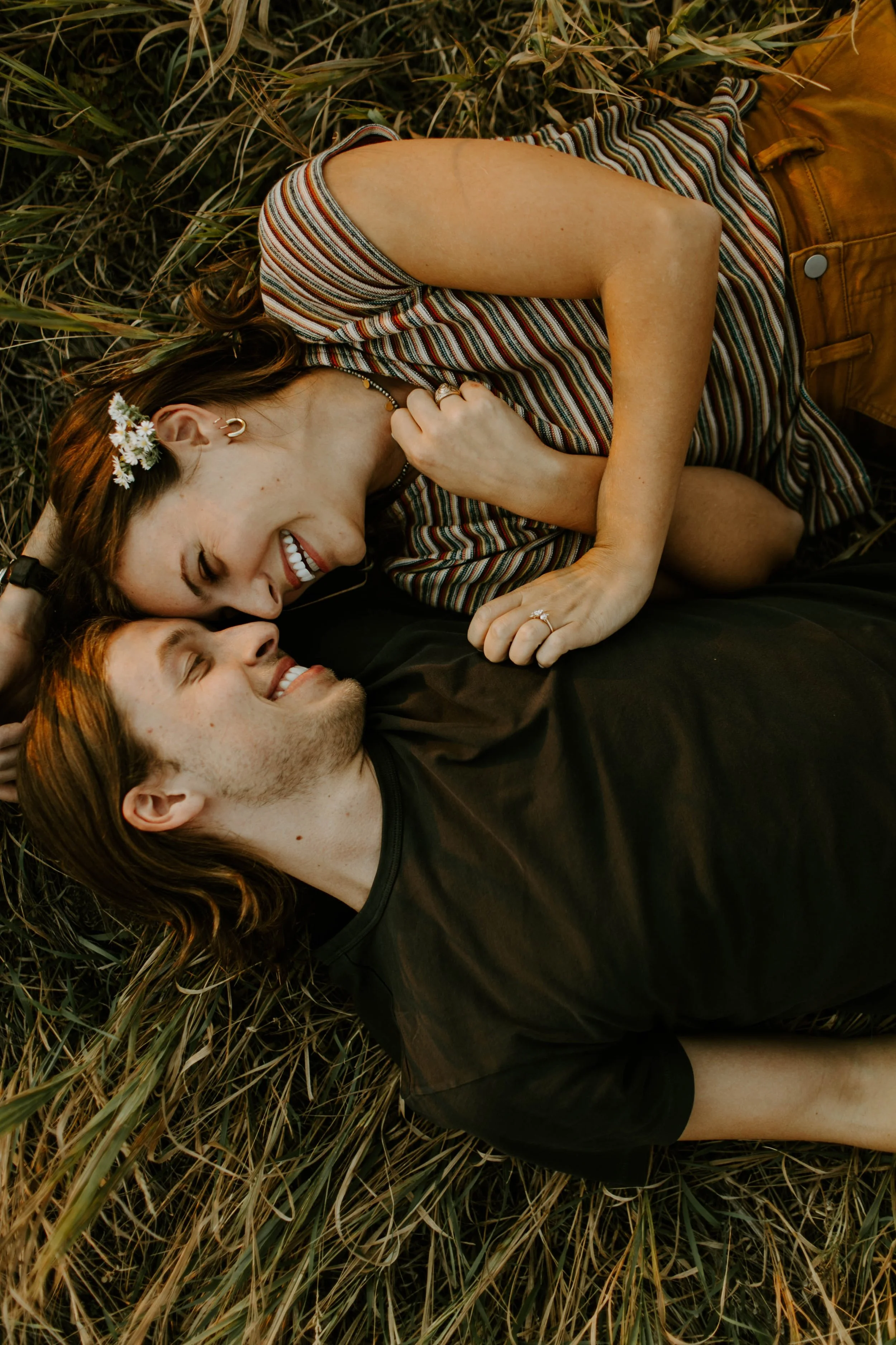 A woman and a man lying on the ground amid grass, smiling and looking into each other's eyes, sharing a loving moment.
