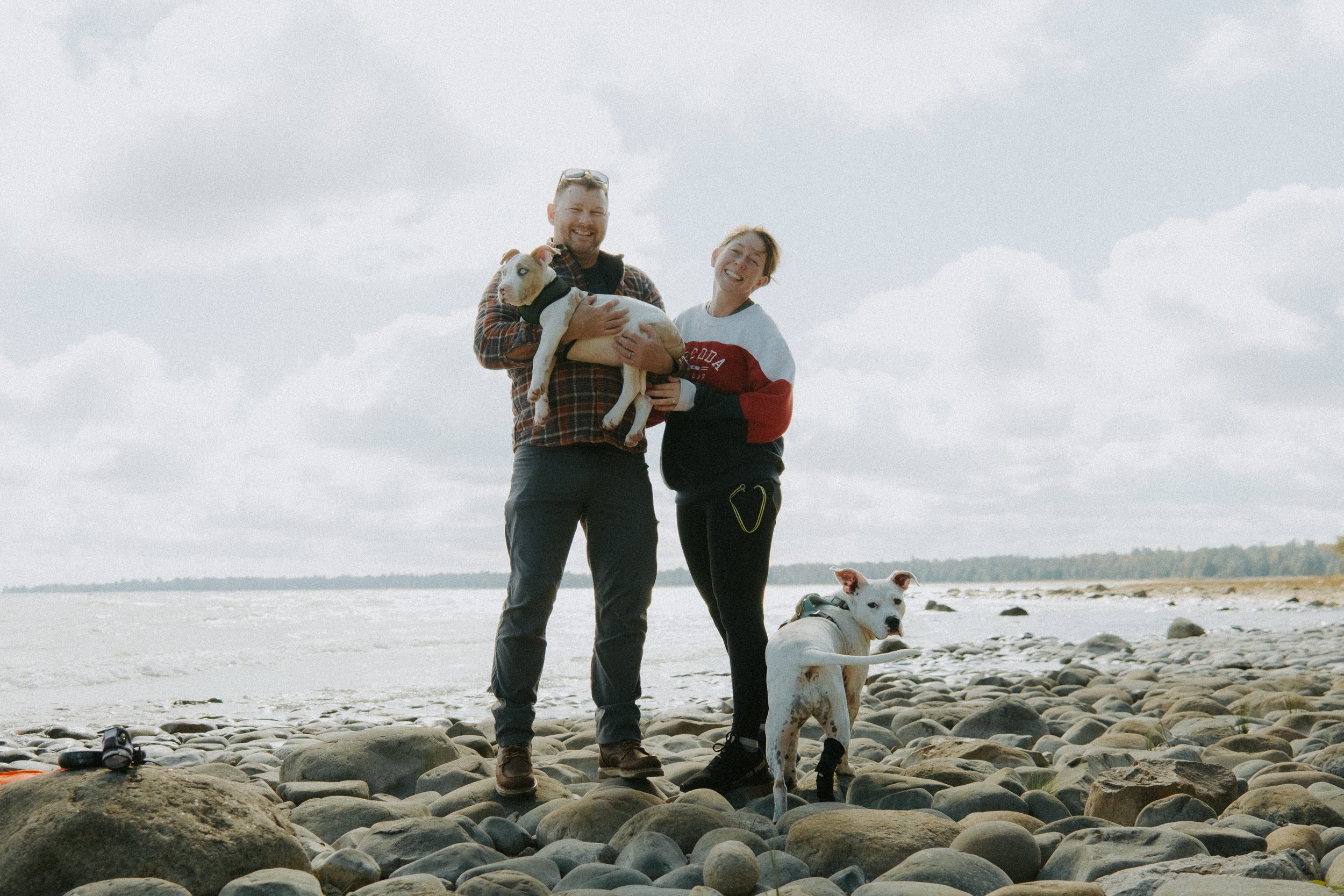 Two people standing on a rocky beach holding a dog, with another dog standing nearby, and the ocean in the background under a cloudy sky.