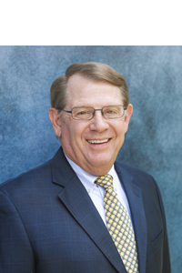 Smiling middle-aged man with glasses and brown hair, wearing a blue checkered shirt, indoors with a blurred modern office background.