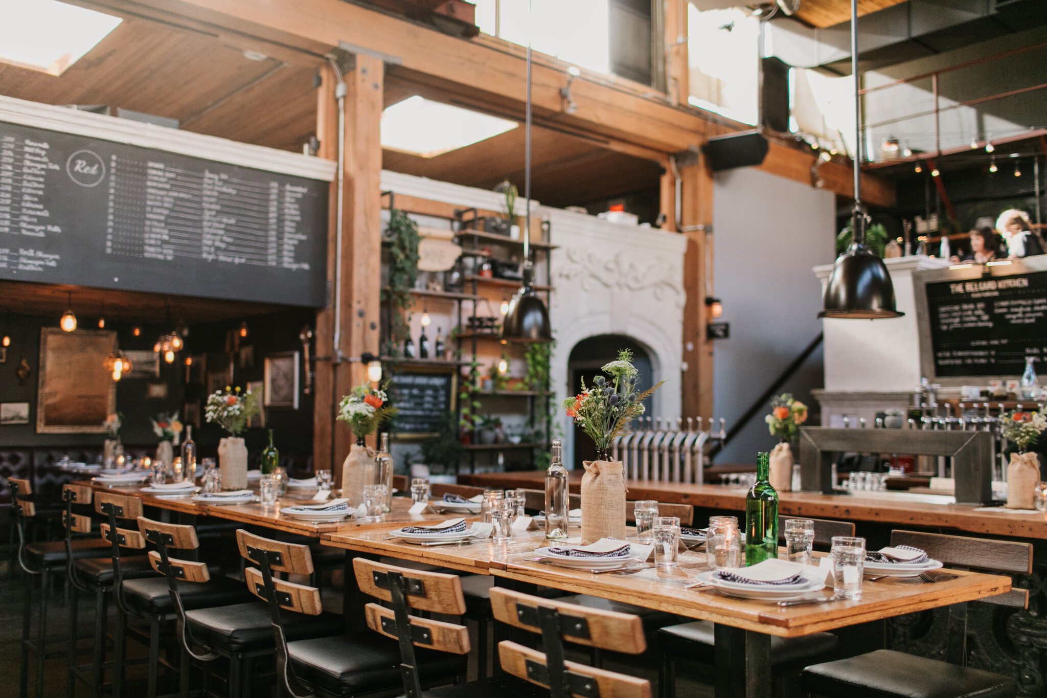 Interior of Belgard Kitchen Vancouver - a rustic restaurant with wooden tables, chairs, and a large menu board. Tables are set with plates, glasses, and flowers in vases. Industrial lighting and decorative elements enhance the cozy ambiance.