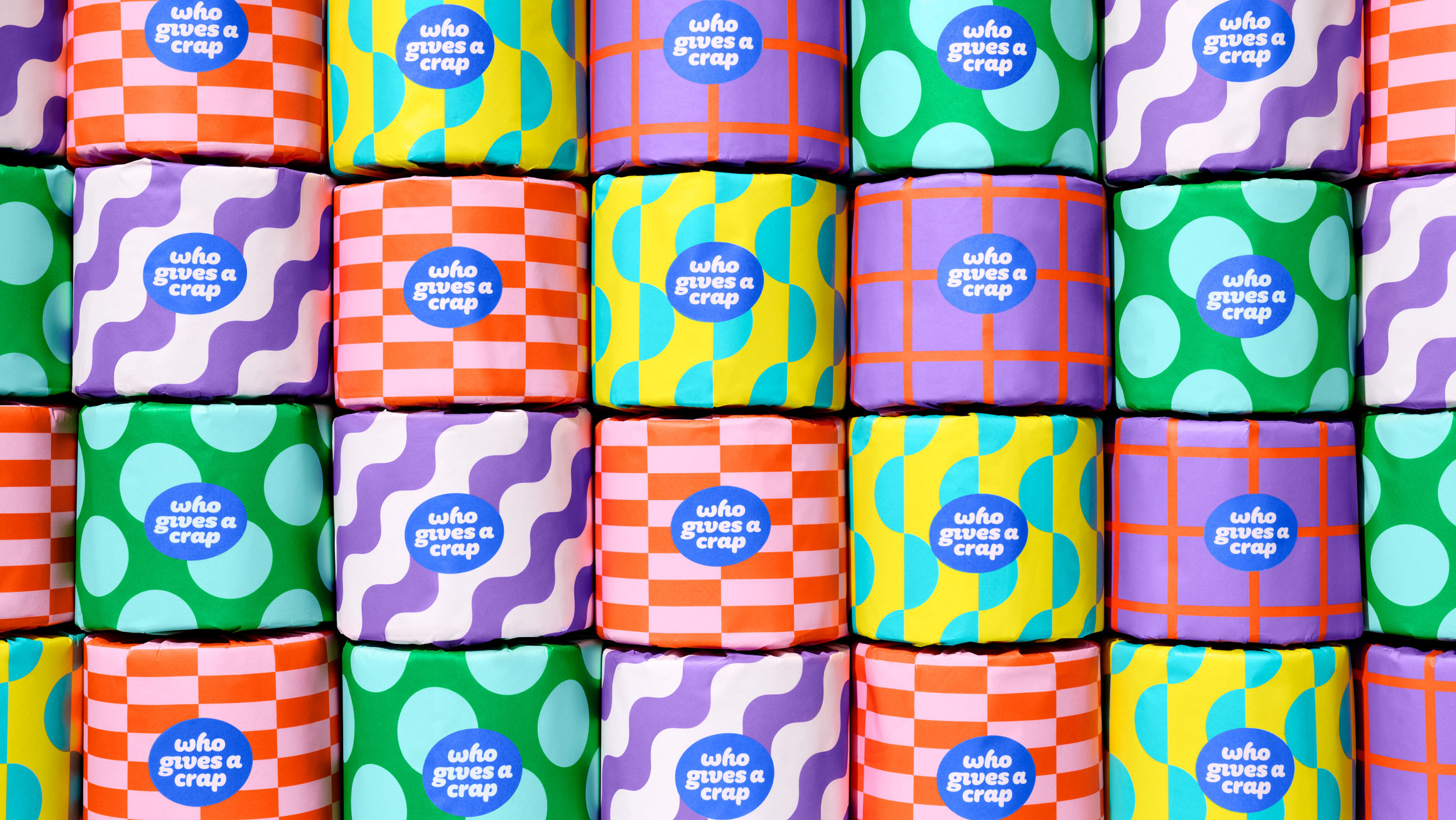 Rolls of Who Gives A Crap toilet paper with vibrant, patterned packaging, stacked together on a bathroom shelf.