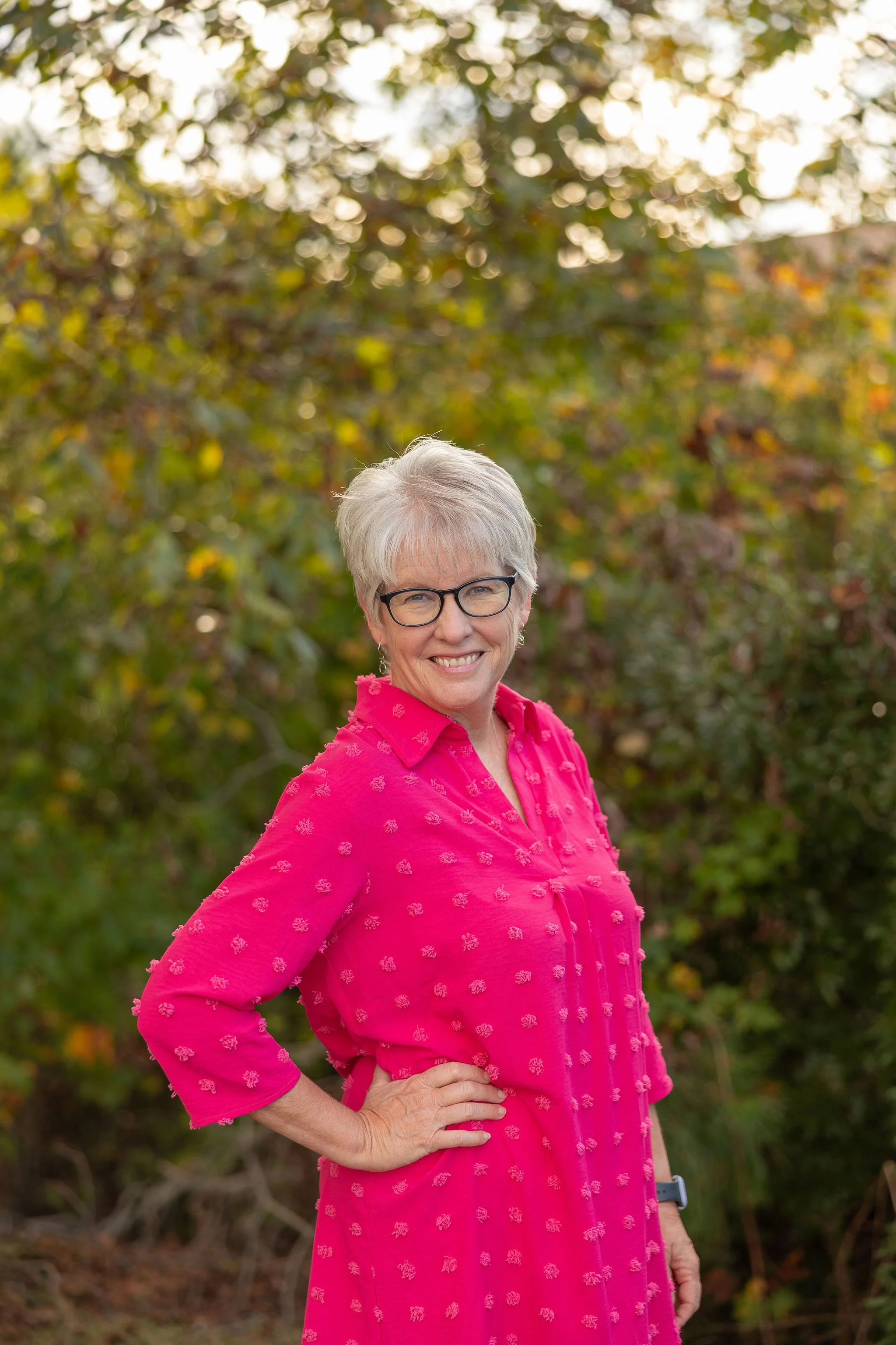 Older woman with short white hair wearing glasses and a bright pink blouse, smiling and standing in front of green foliage.