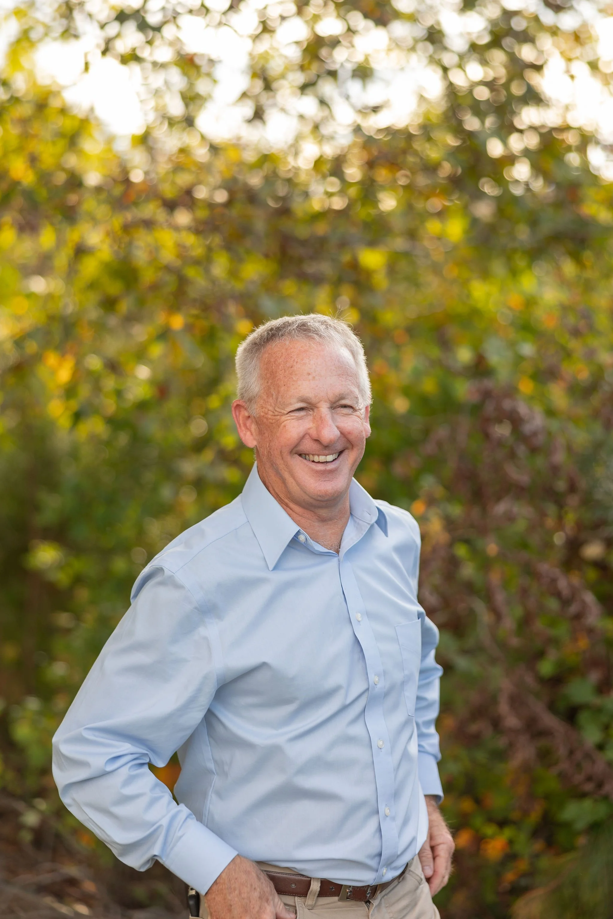 Smiling man in a light blue shirt in front of a blurred green foliage background.