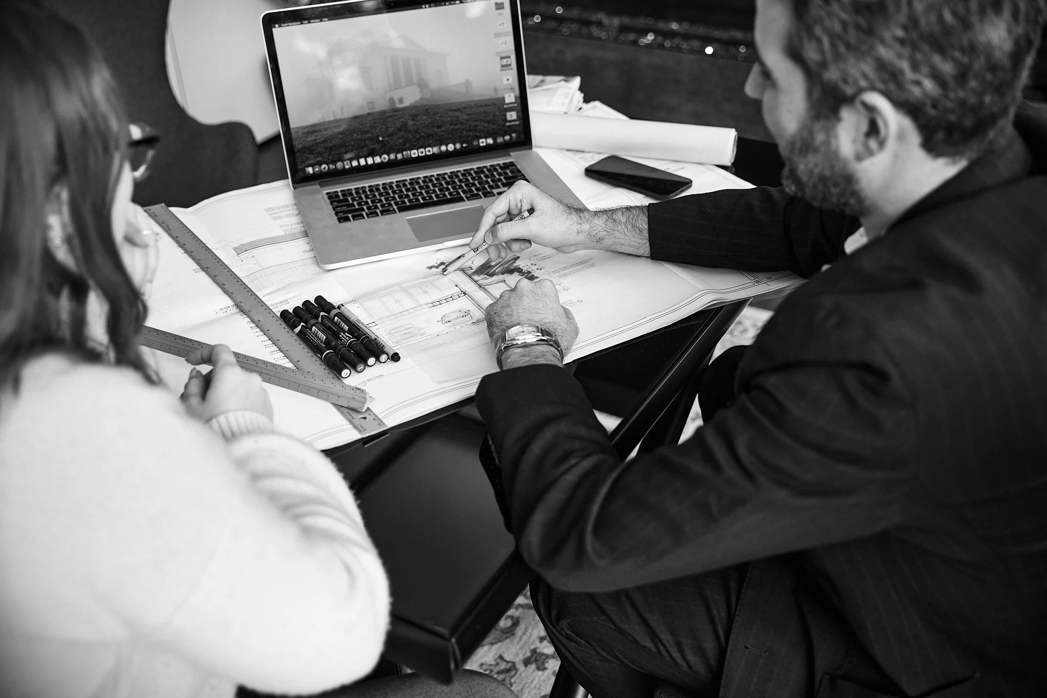 Two people working together at a desk with a laptop, architectural plans, rulers, pens, and a smartphone in black and white.