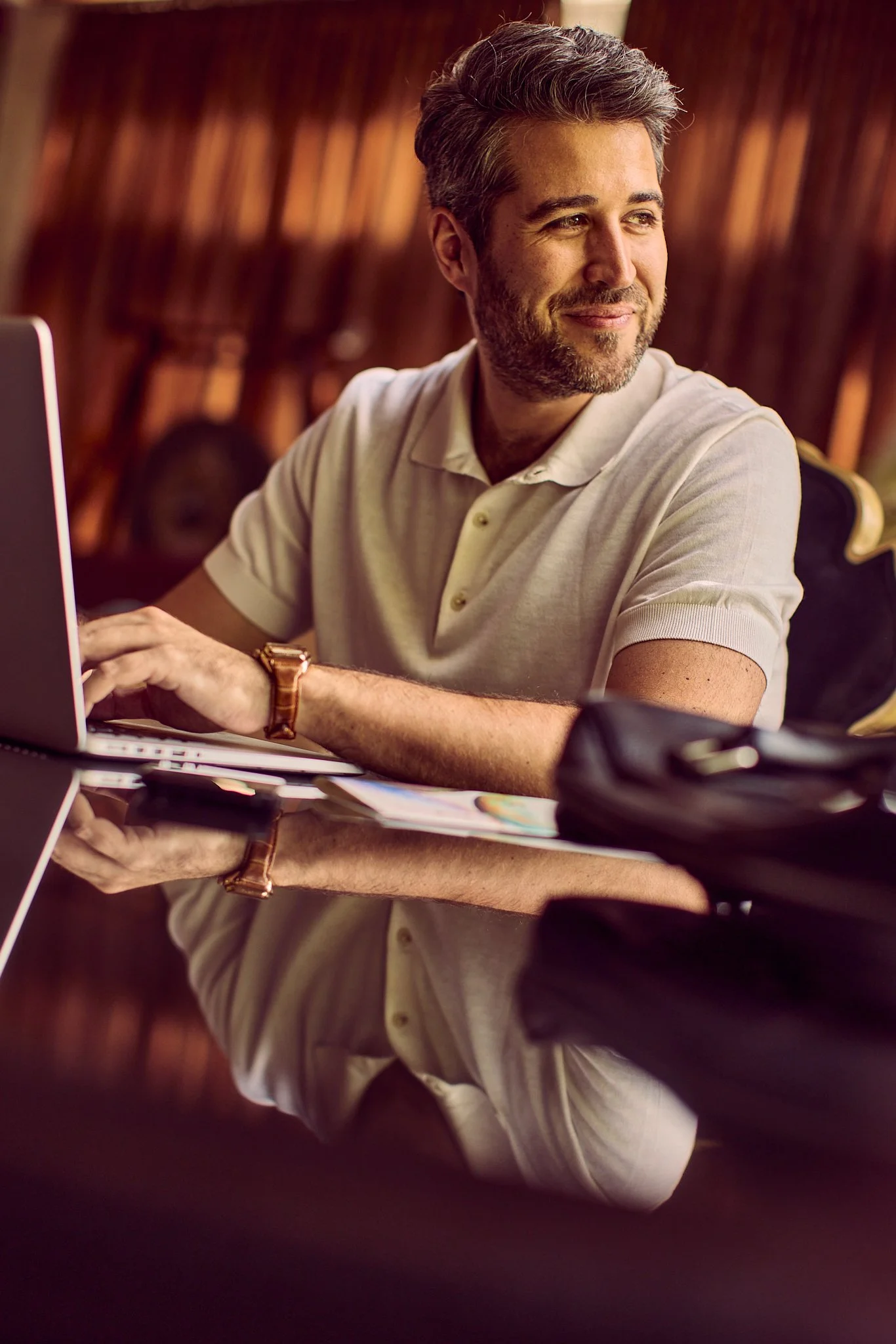 A man sitting at a polished wooden table with a laptop, smiling and looking to the side, in a warmly lit room.