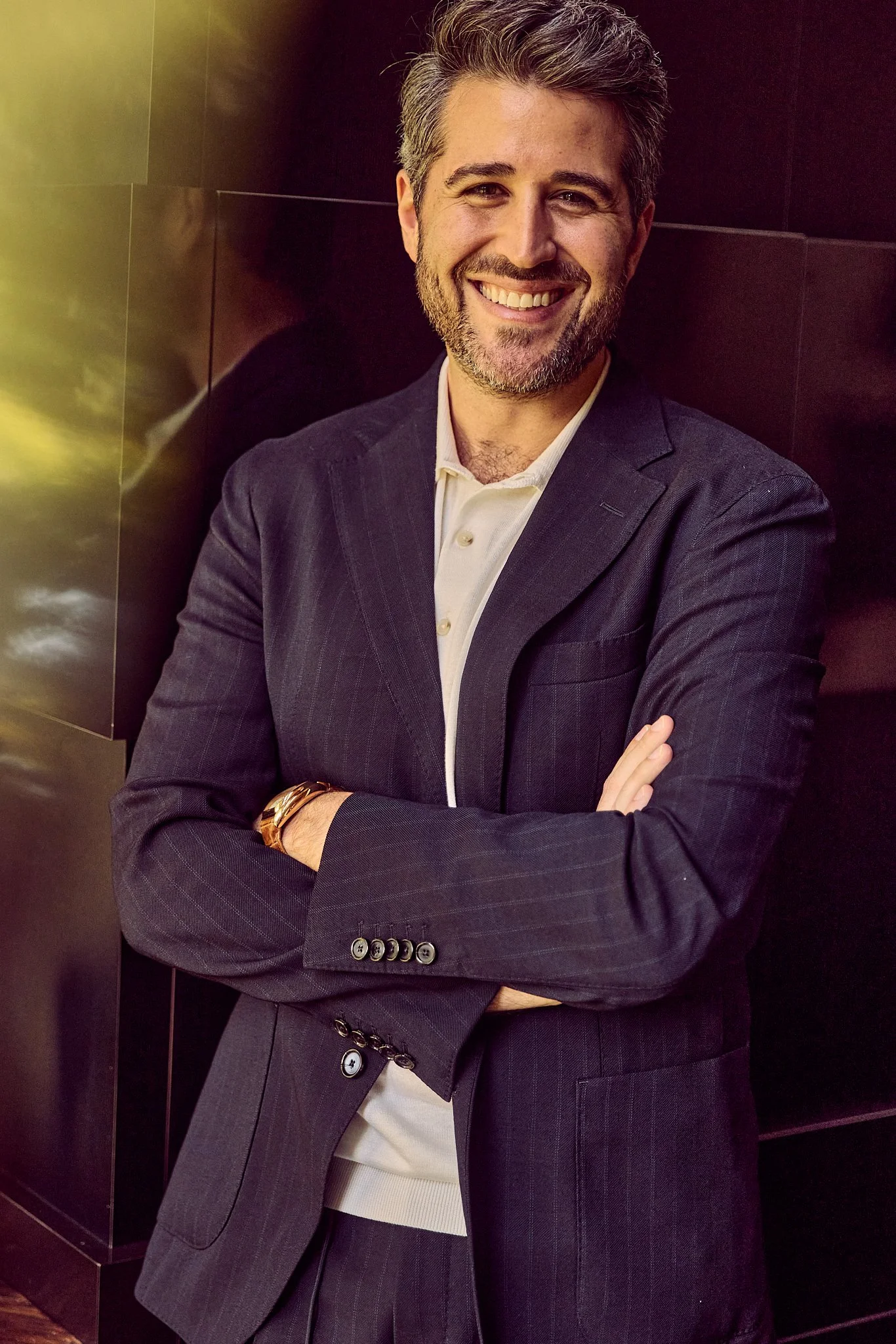 A smiling man in a dark pinstripe suit with arms crossed, standing against a dark background with a gold and green hue, looking at the camera.