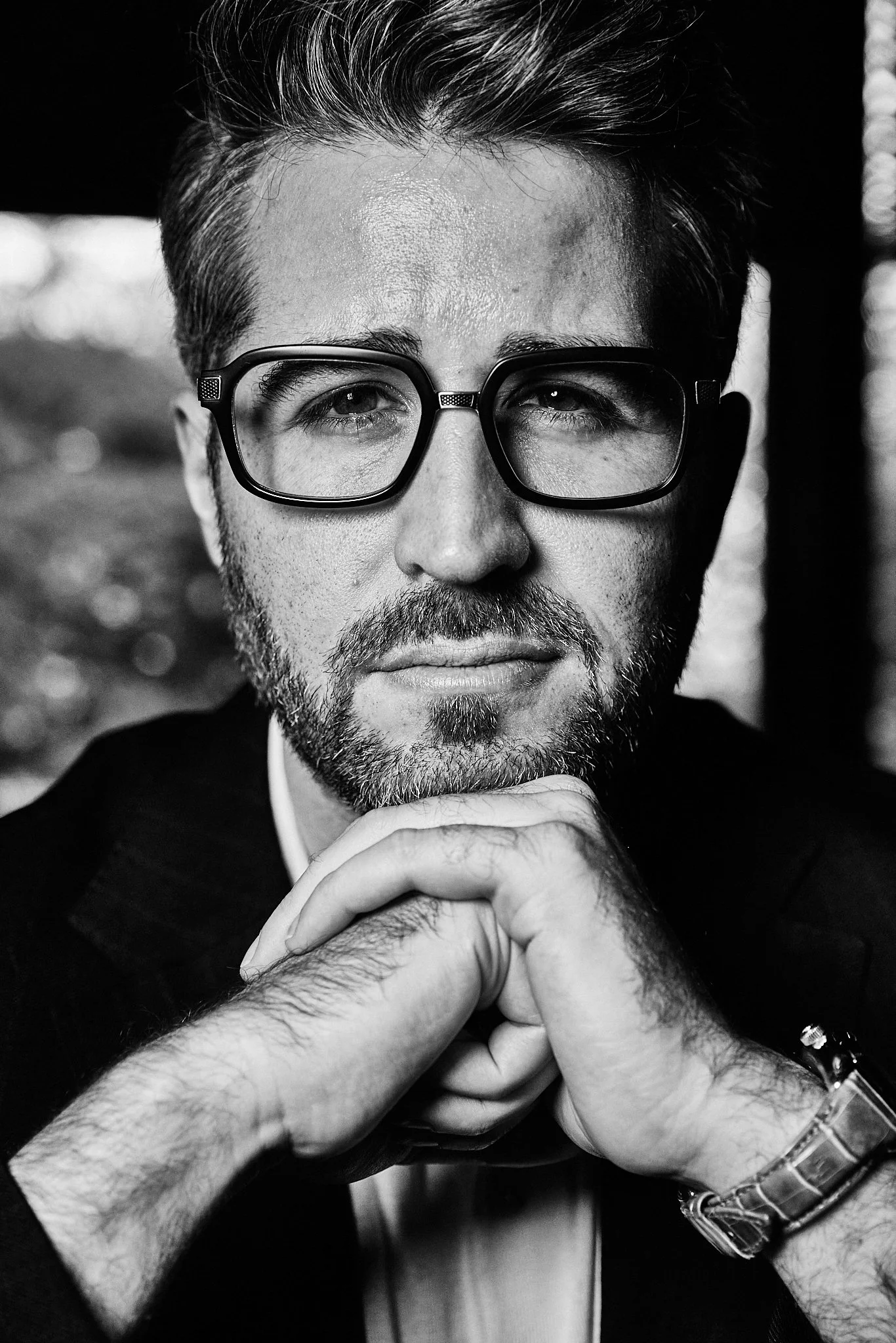 A black-and-white close-up portrait of a man with short hair, beard, glasses, and a serious expression, resting his chin on his hands.