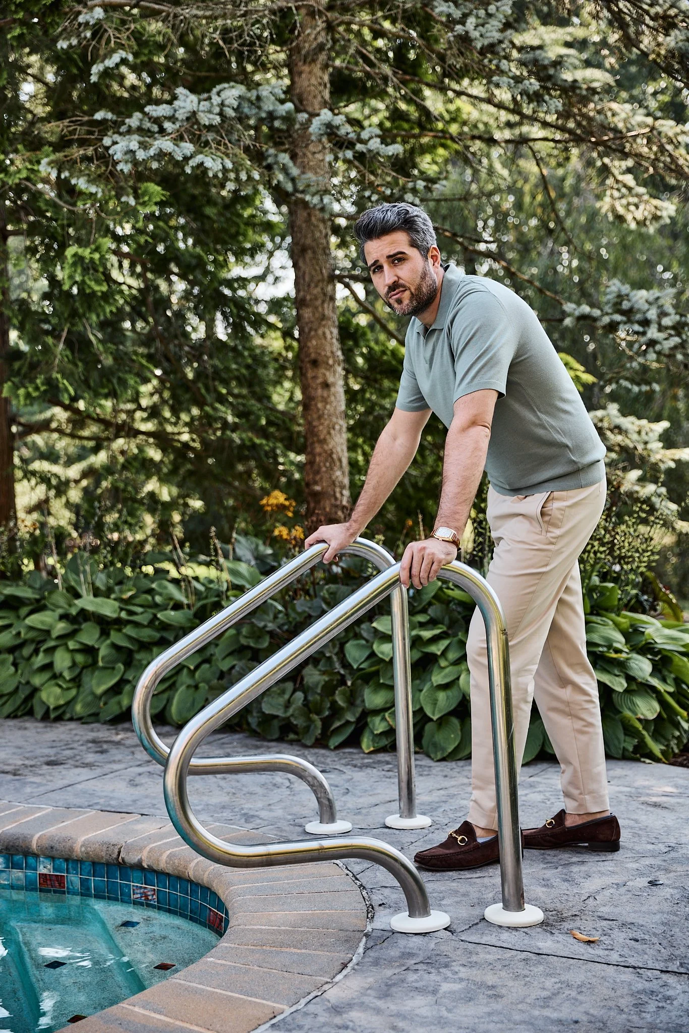 A man with gray hair and a beard gets into a swimming pool, holding onto the metal handrails at the pool's edge, surrounded by lush green plants and trees.