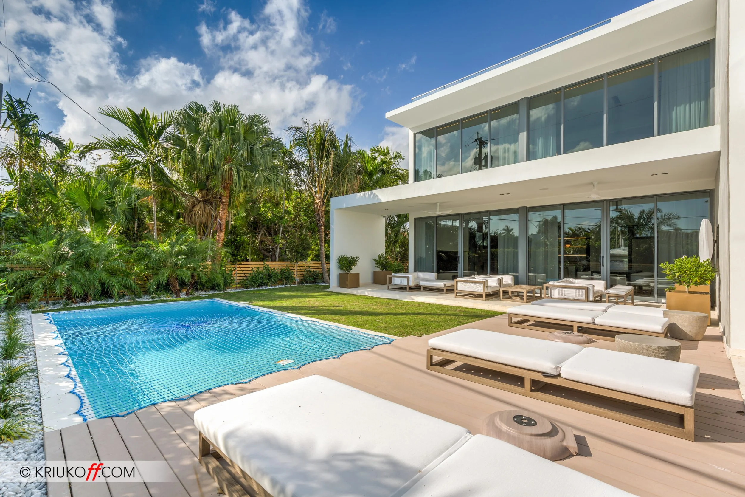 Modern house with large glass windows, outdoor lounge seating, and a small pool surrounded by greenery and palm trees under a partly cloudy sky.