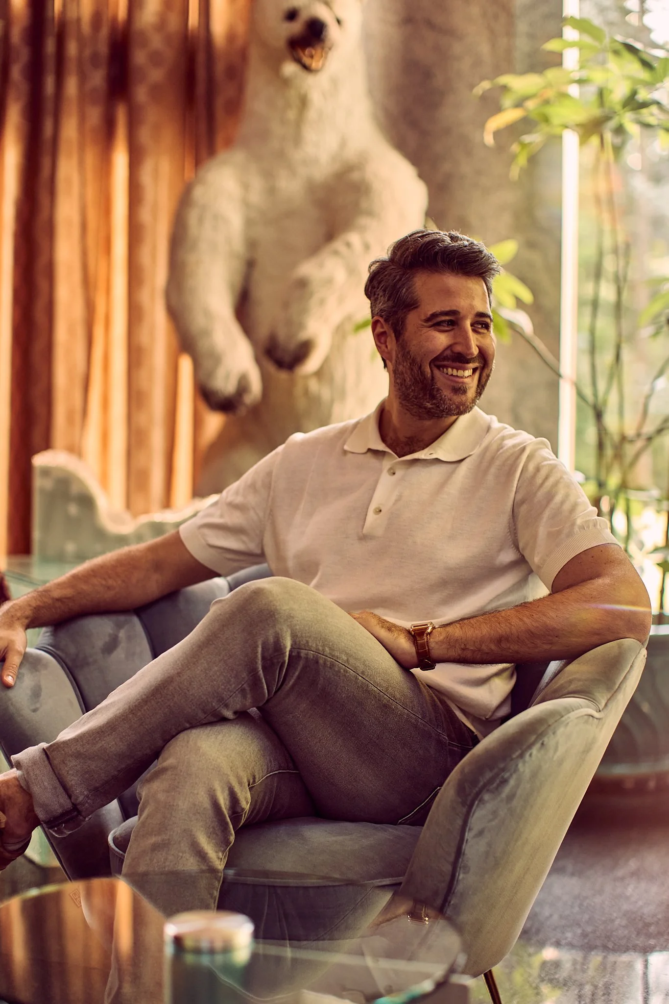 A man sitting on a blue armchair, smiling, in a room with a large bear statue in the background and plants near the window.