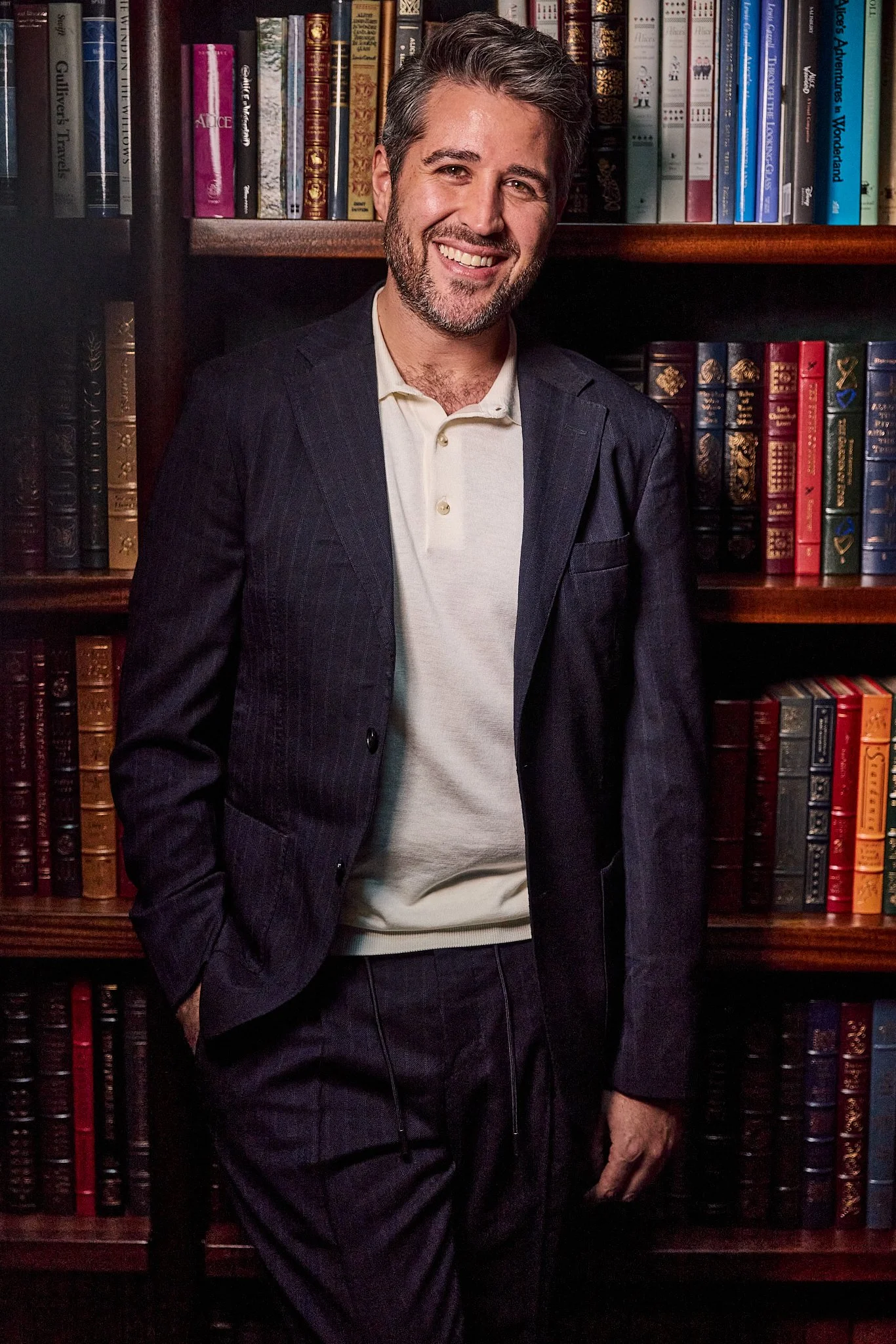 A man in a dark suit and light-colored shirt stands in front of a bookshelf filled with colorful books.