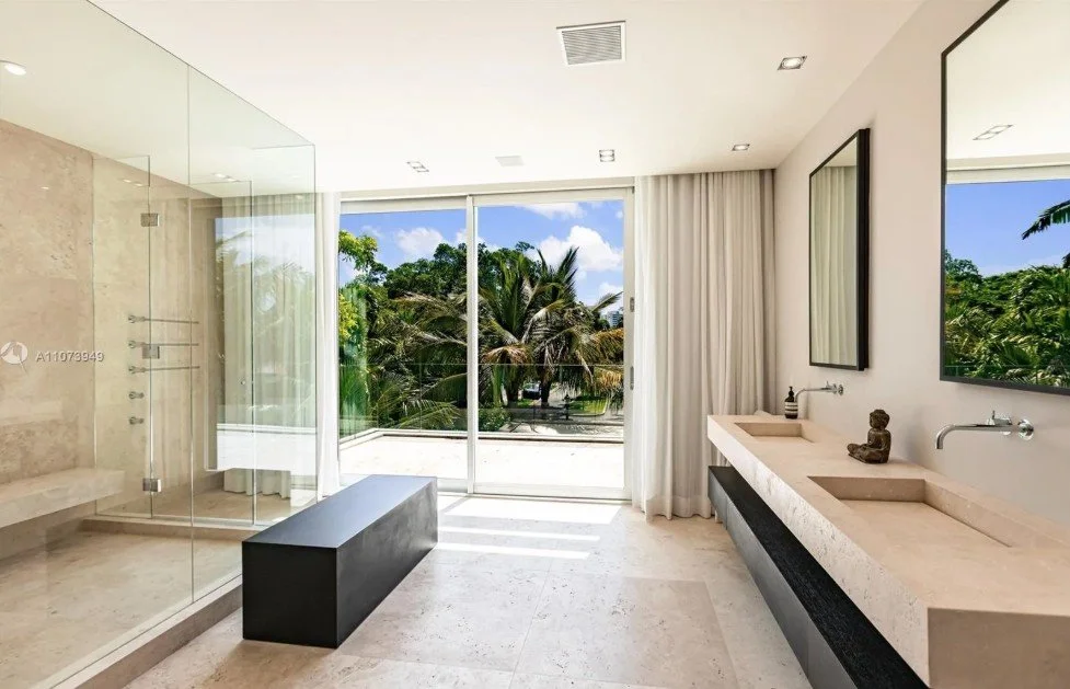 the primary bathroom with travertine flooring, floor to ceiling glass, modern fixtures, flos lighting, and palm trees beyond.