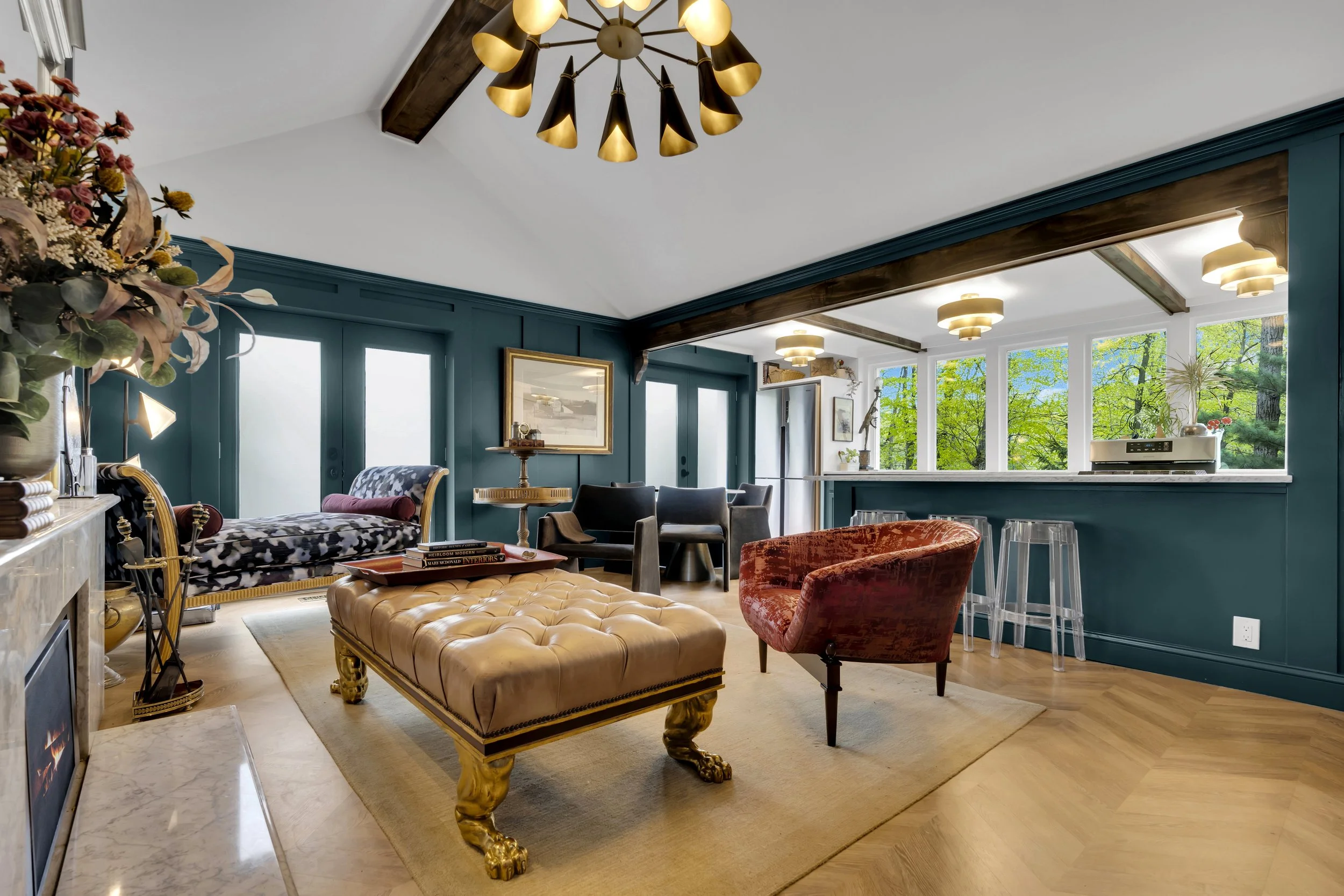 Living room with dark teal walls, large corner windows showing green trees outside, a gold-trimmed beige tufted leather ottoman with lion paw feet, a red velvet armchair, a black armchair, and a patterned chaise lounge. Decor includes a large gold-framed mirror, books, lamps, and a chandelier with black and gold shades.