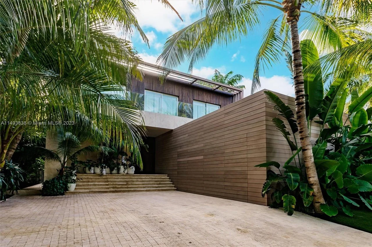 Modern house entrance with stairs, surrounded by tropical palm trees and lush greenery, under a partly cloudy sky.