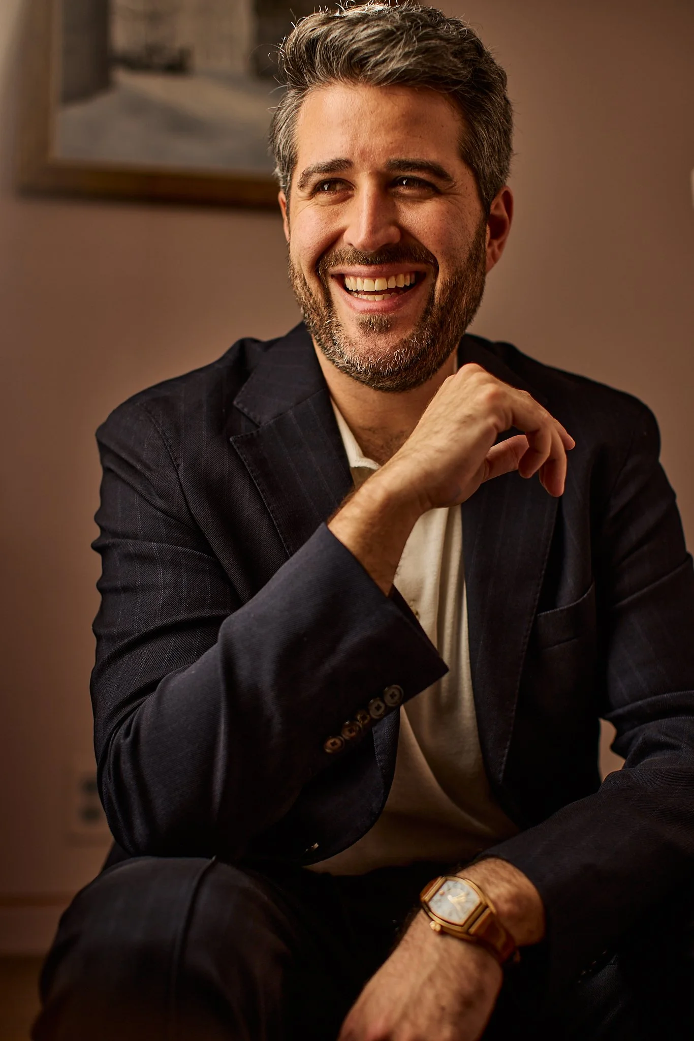 A middle-aged man with gray hair and a beard, smiling while sitting indoors. He is wearing a dark blazer, a white shirt, and a gold watch on his left wrist.
