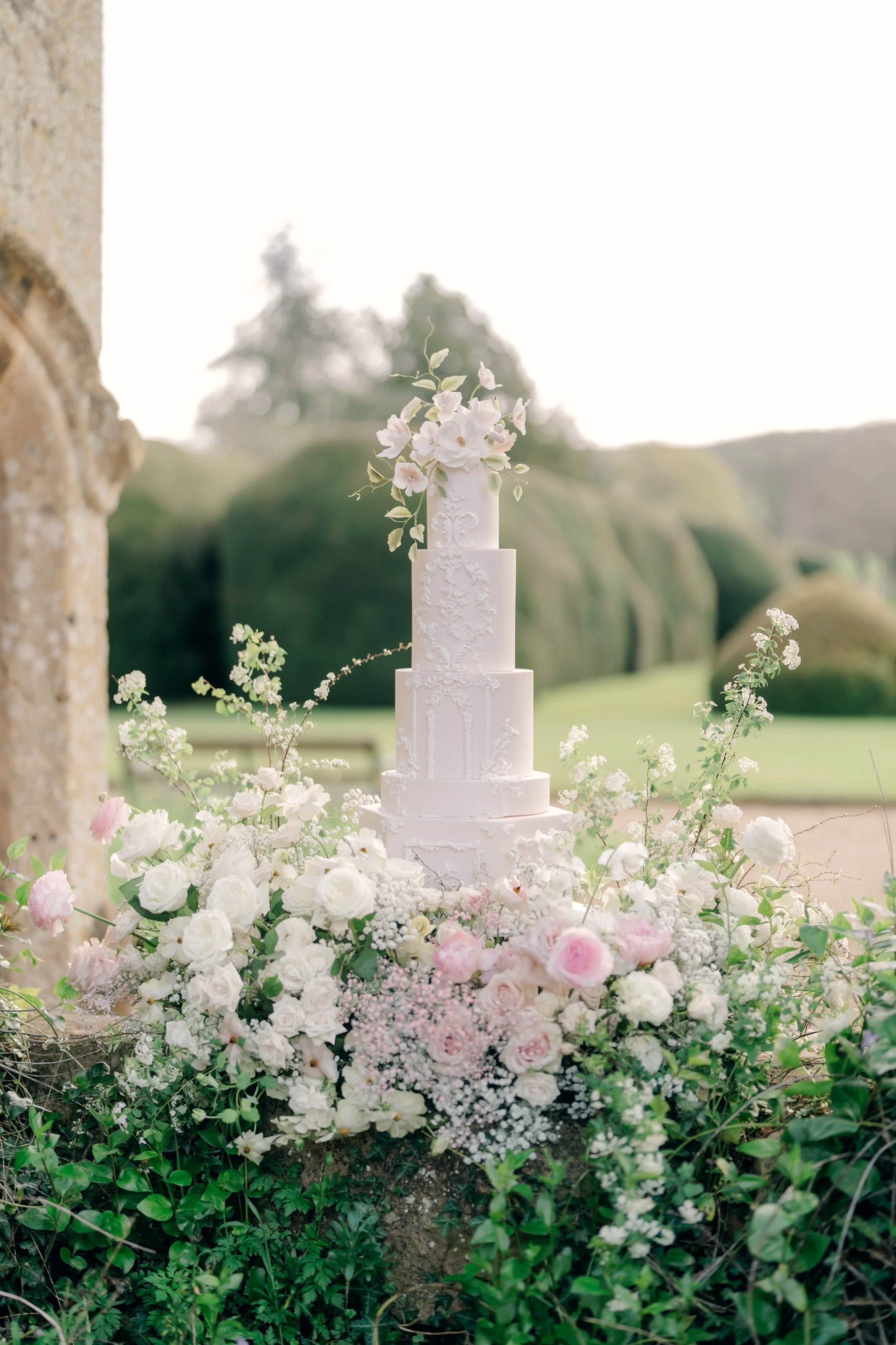 A tall, white wedding cake decorated with intricate patterns, topped with white and pink flowers, and surrounded by white and pink roses and greenery, set outdoors on a sunny day.
