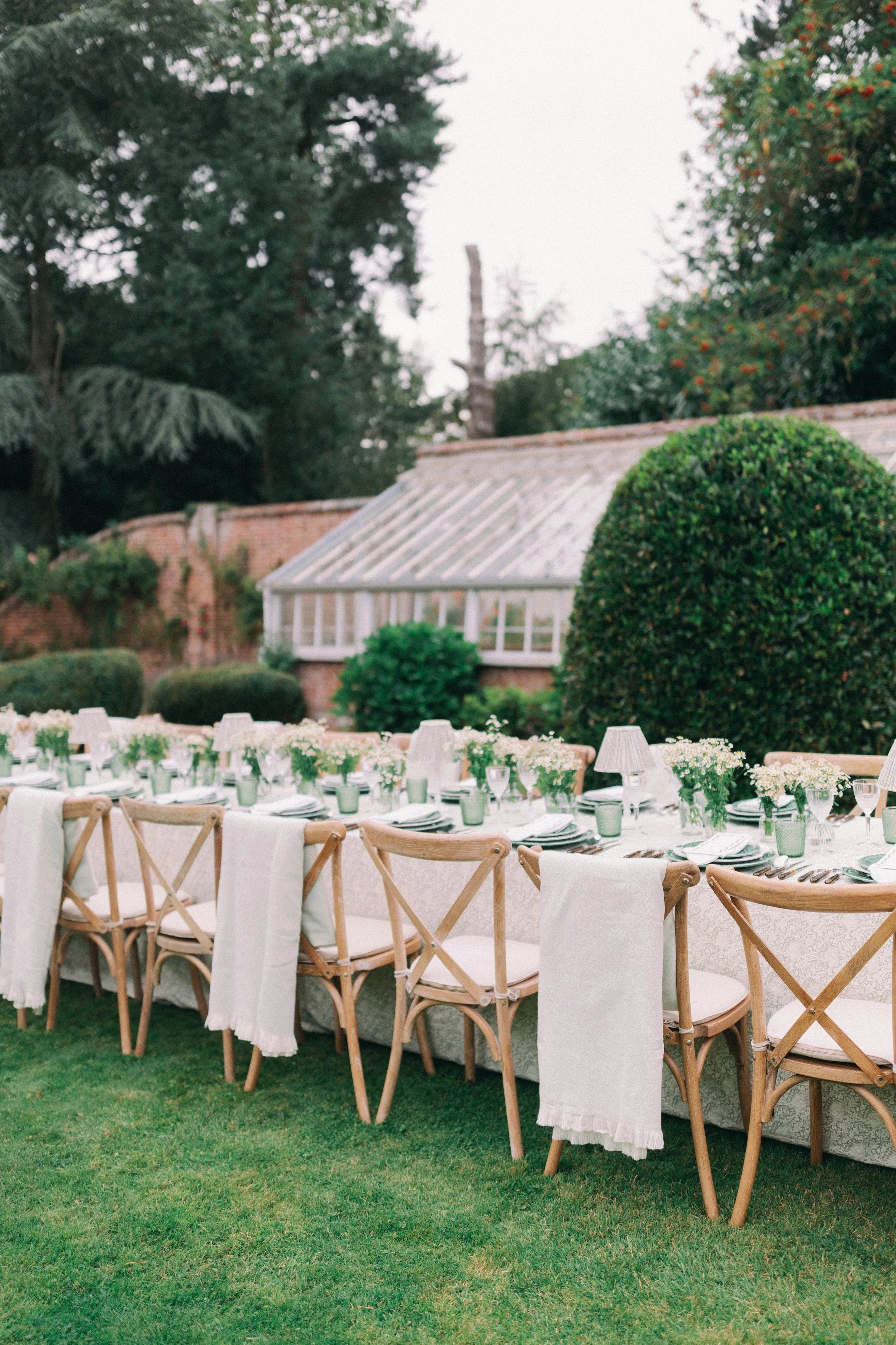 Outdoor garden table set for a formal event with floral tablecloth, floral bud vases, tableware, and wooden chairs, surrounded by lush greenery and a greenhouse. Hosted In Norfolk near Holt