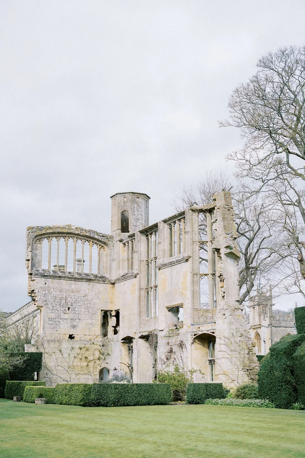 Ruins of an old stone building with large windows and a tower, surrounded by green grass, bushes, and leafless trees under a cloudy sky.