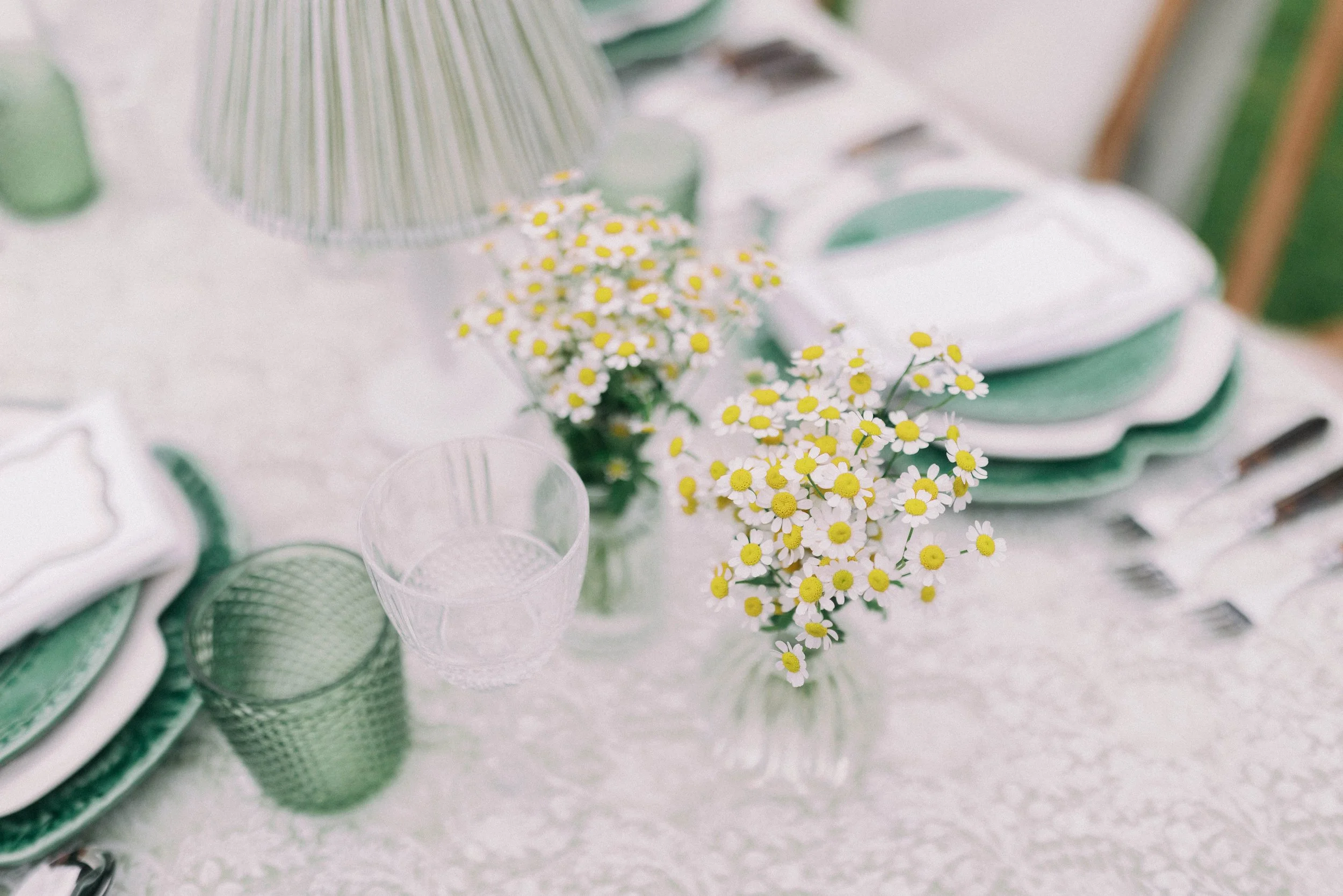 Table setting with a bud vases of daisies, green and white dinnerware, and drinking glasses on a floral tablecloth. Hosted near Holt in Norfolk