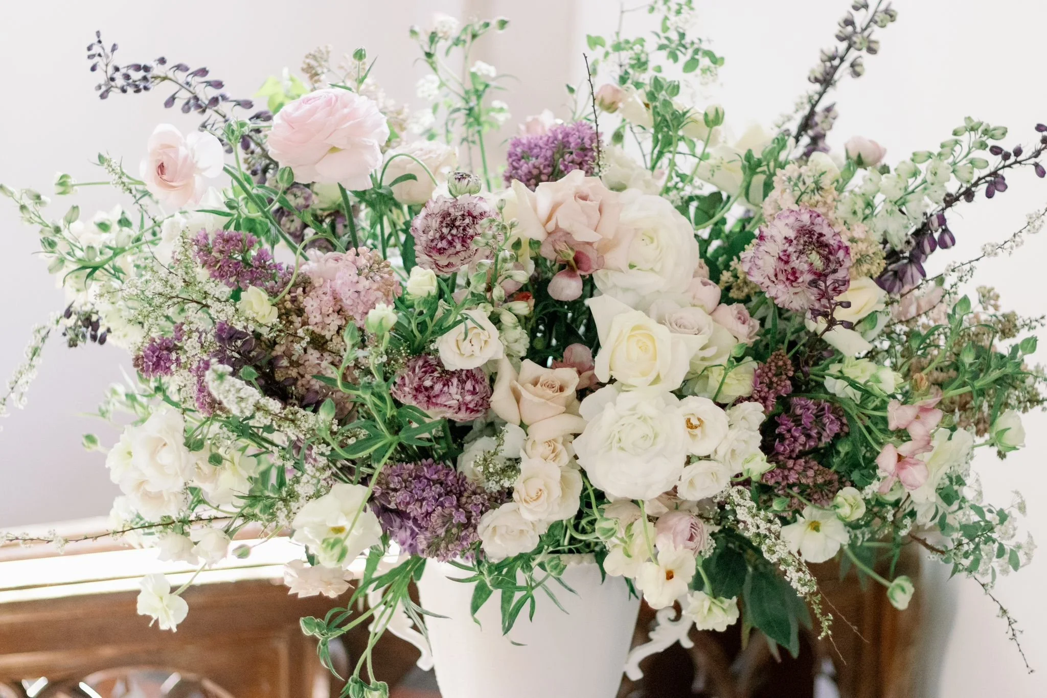A large arrangement of mixed flowers including white roses, purple lilacs, and light pink blossoms in a white vase on a wooden surface.