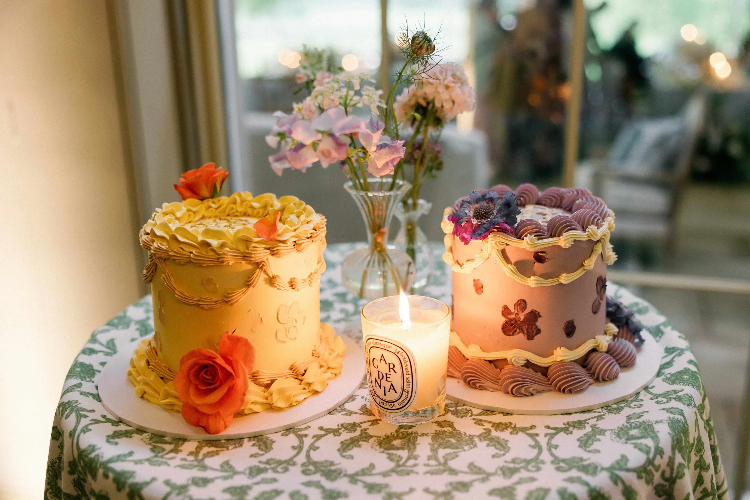 Two decorated cakes with flowers on top, placed on a table with a floral tablecloth, and a lit candle between them, in a room with a glass door and some furniture in the background.