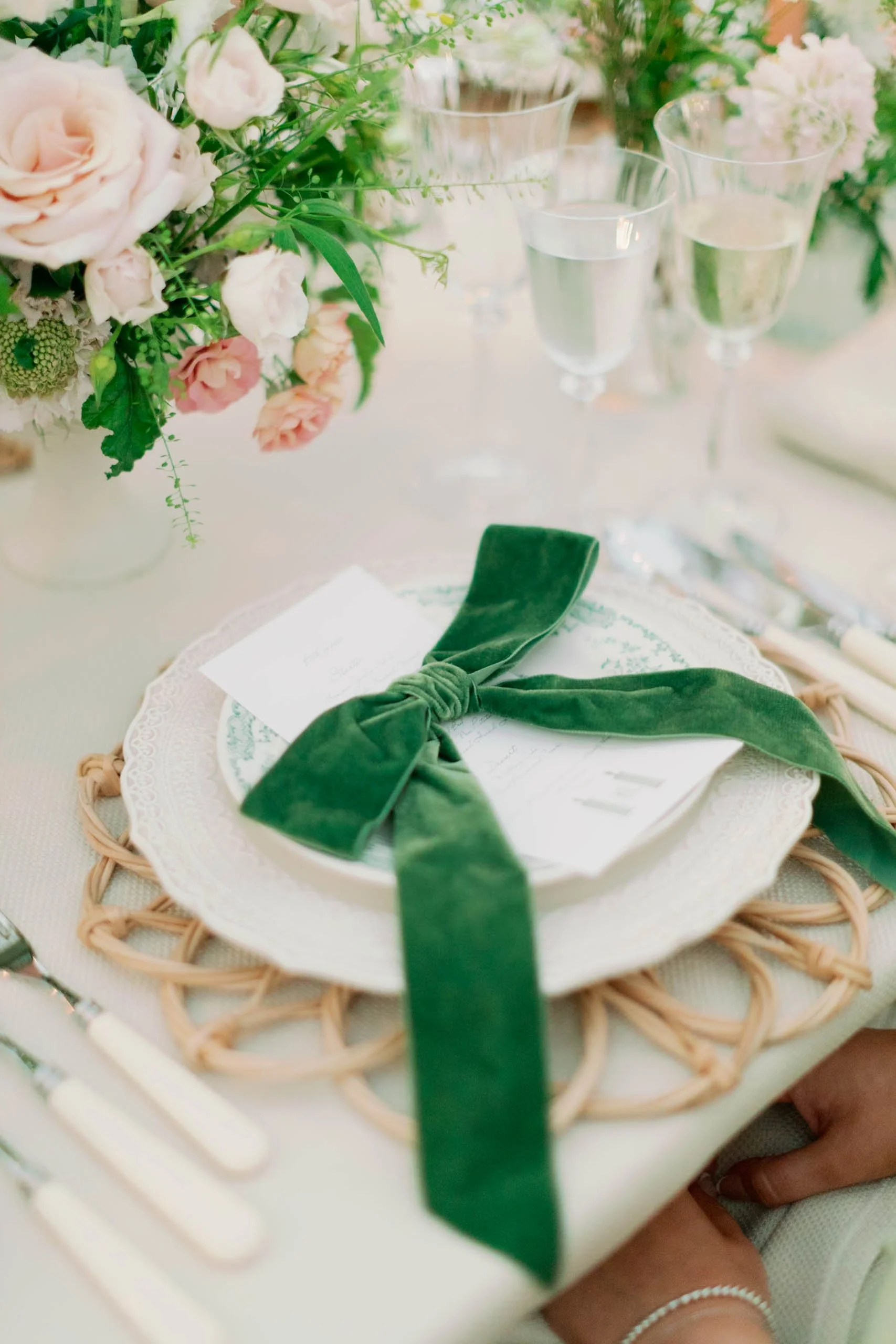 Elegant table setting with a green velvet bow tied around a menu on white plates, with glassware and floral centerpieces.