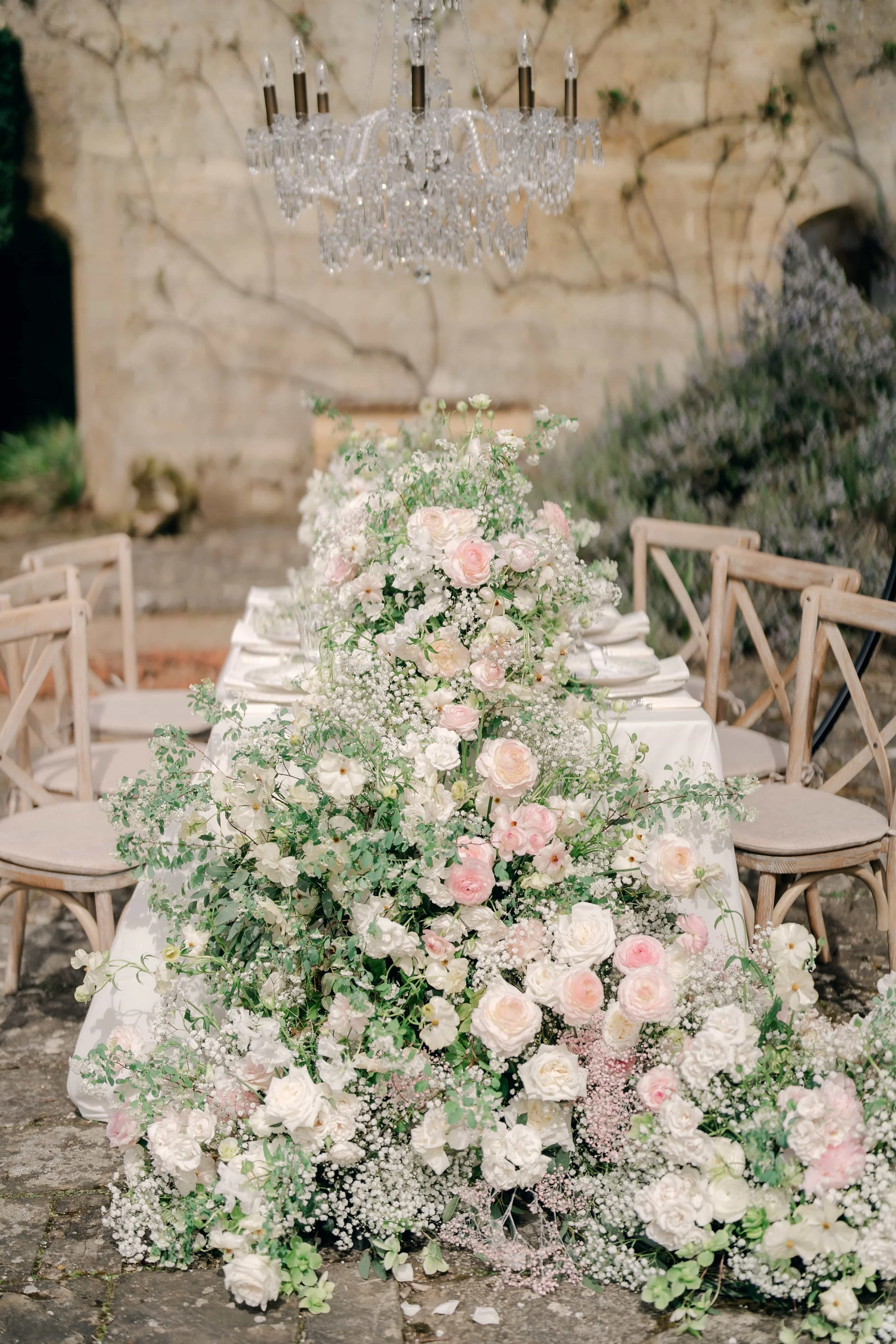 A long floral centerpiece with white and pink roses, baby's breath, and greenery on a dining table, with a crystal chandelier hanging above, in an outdoor setting with stone walls and wooden chairs.
