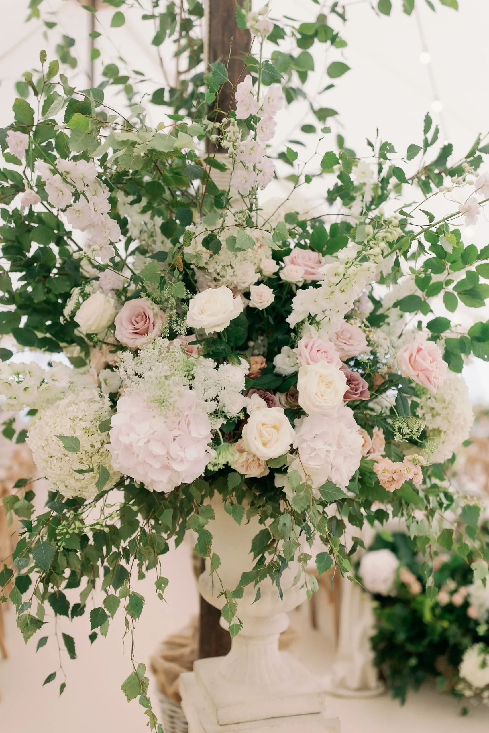 A large floral arrangement in a white urn contains white and light pink roses, white hydrangeas, and various green foliage.