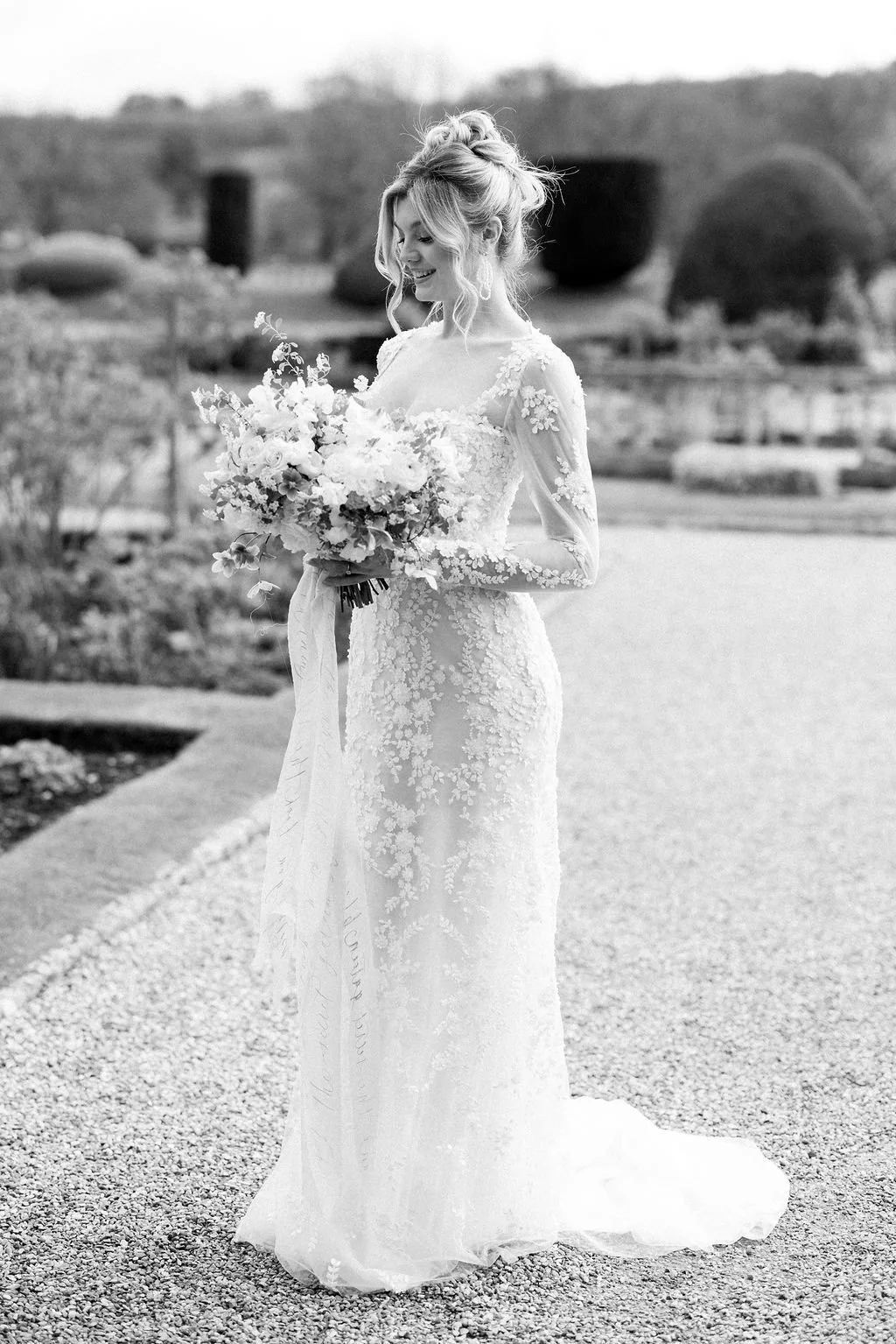 Black and white photo of a joyful bride in a long lace wedding dress, holding a large bouquet of flowers, standing outdoors on a gravel path.