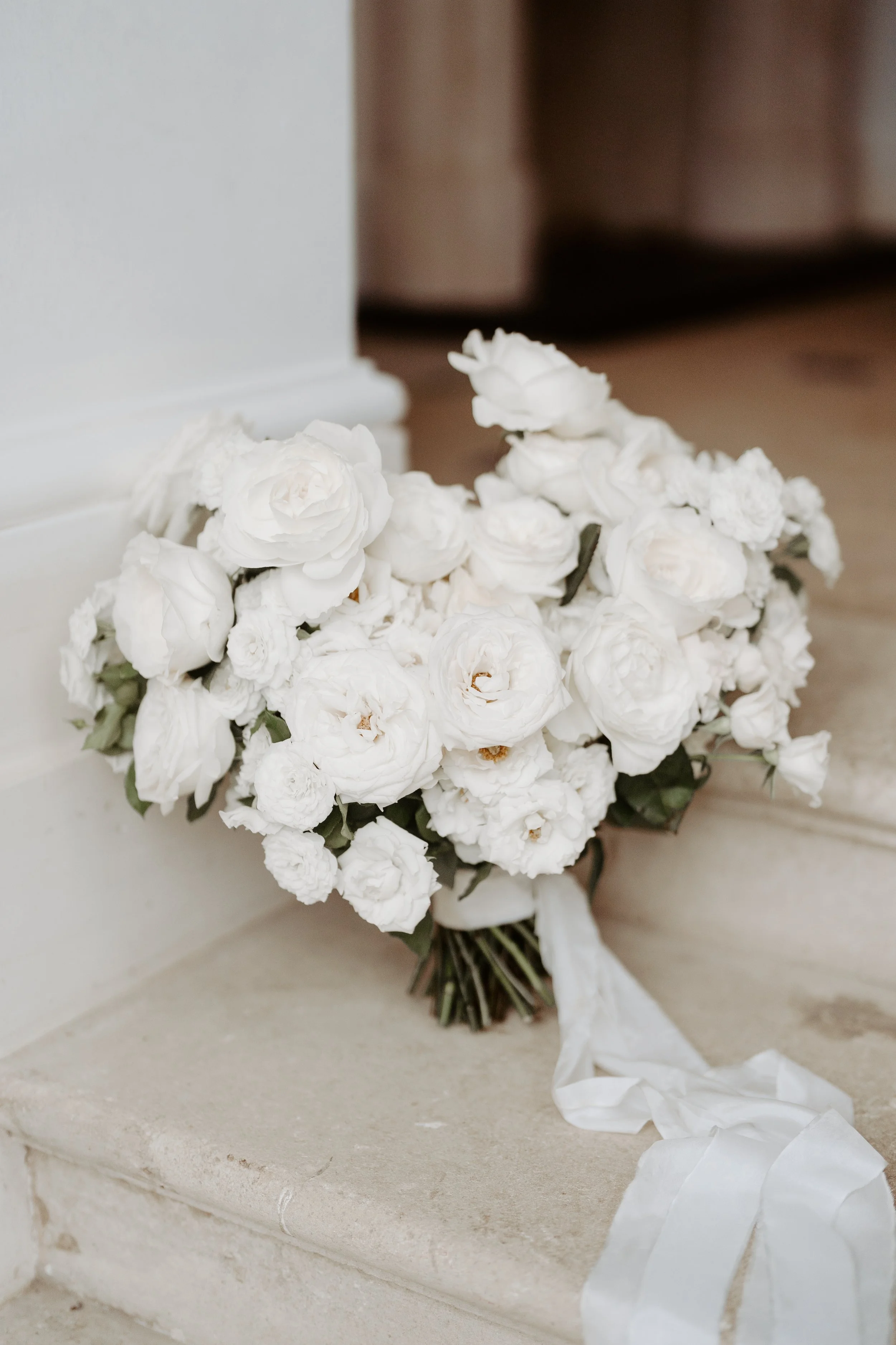 A bouquet of white flowers, likely roses or peonies, placed on a light-colored stone step near a white wall and wooden floor, tied with a white ribbon.