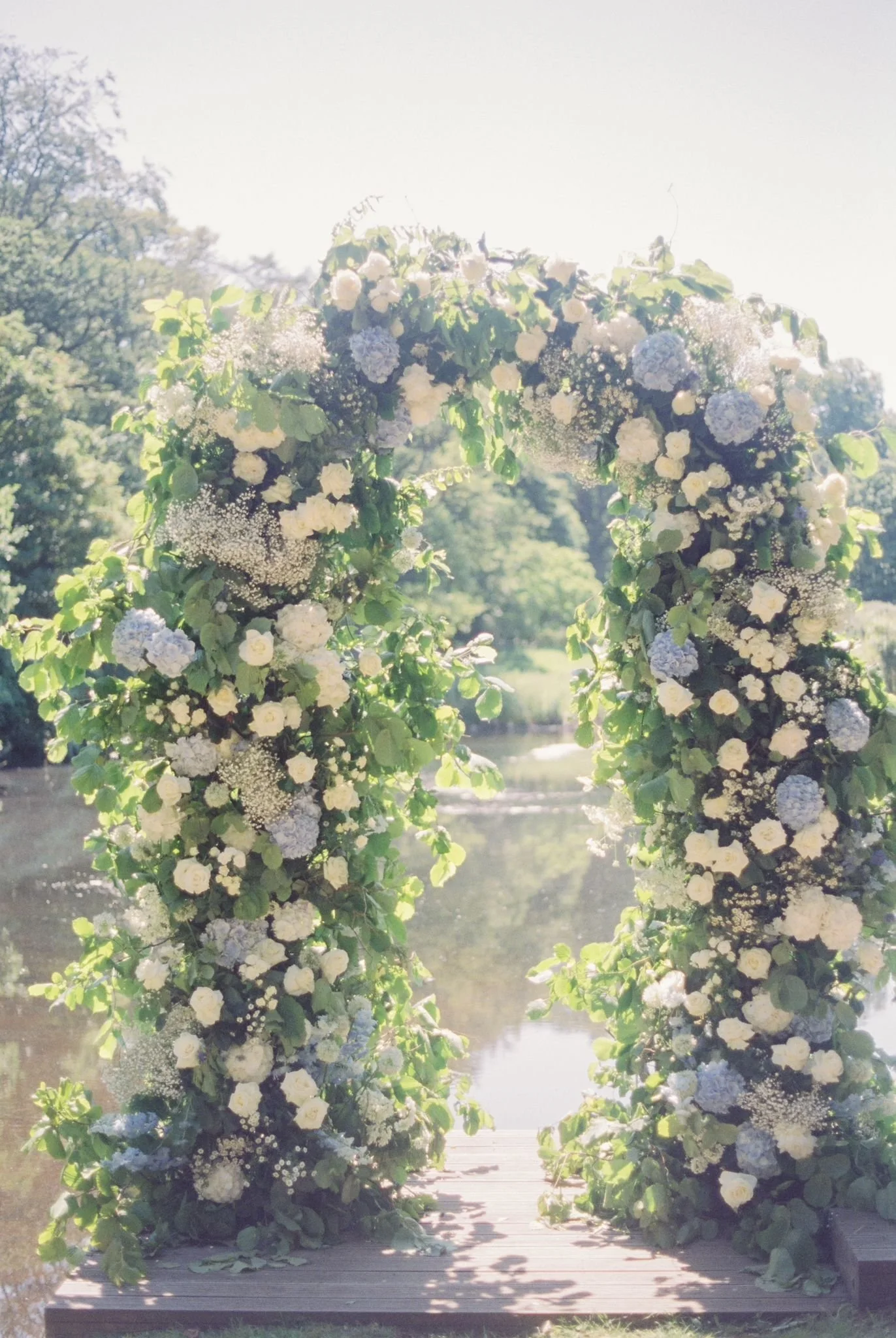 A floral wedding arch decorated with white and blue flowers on a wooden platform by a river, surrounded by trees, in bright sunlight.