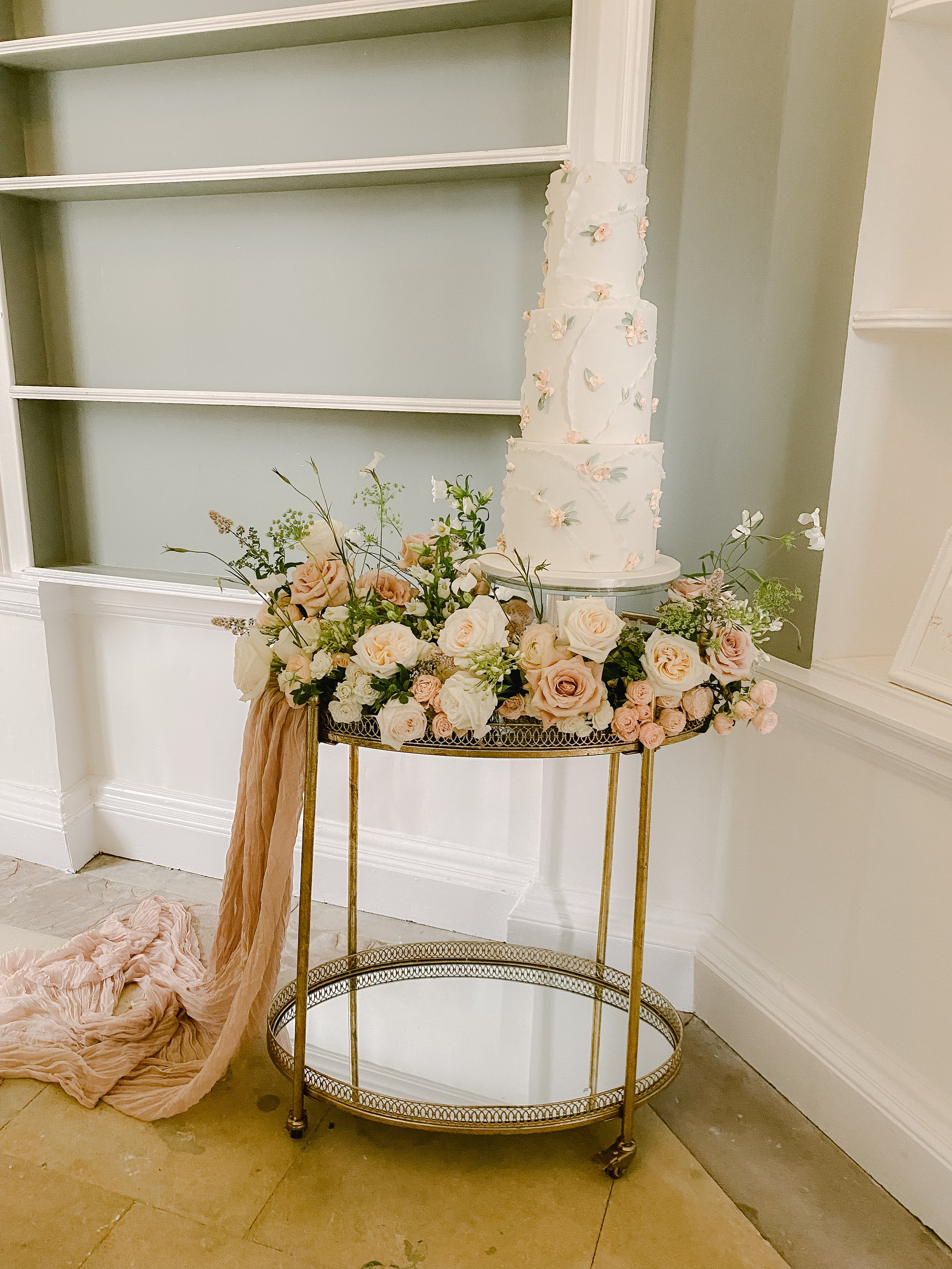 A tall, four-tiered white wedding cake with pastel floral decorations, set on a glass and gold stand surrounded by a large arrangement of pastel roses and greenery on a gold table. There is a soft pink fabric hanging over the side of the table.