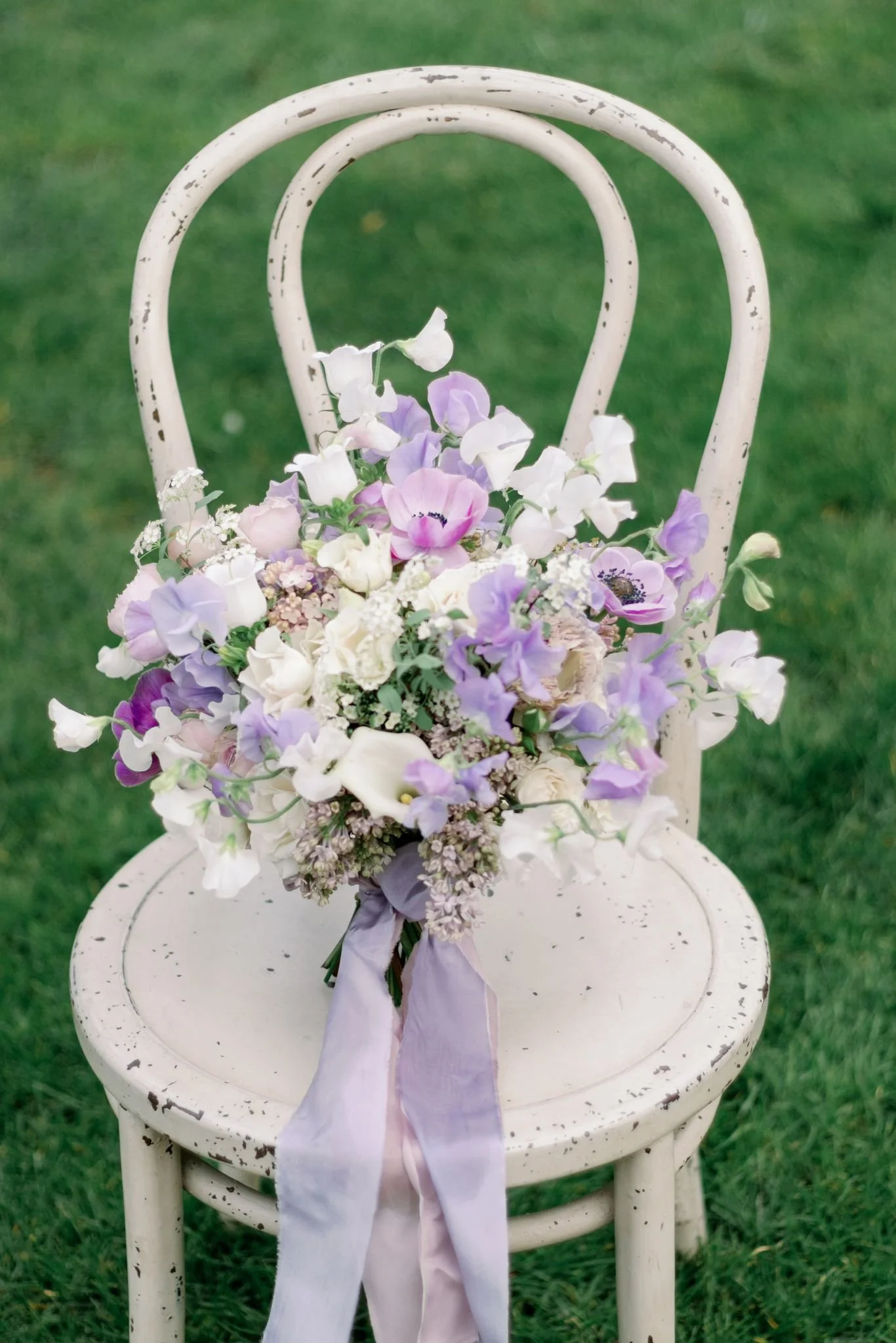A bouquet of purple, white, and cream flowers tied with a purple ribbon, placed on a vintage white distressed wooden chair outdoors on green grass.