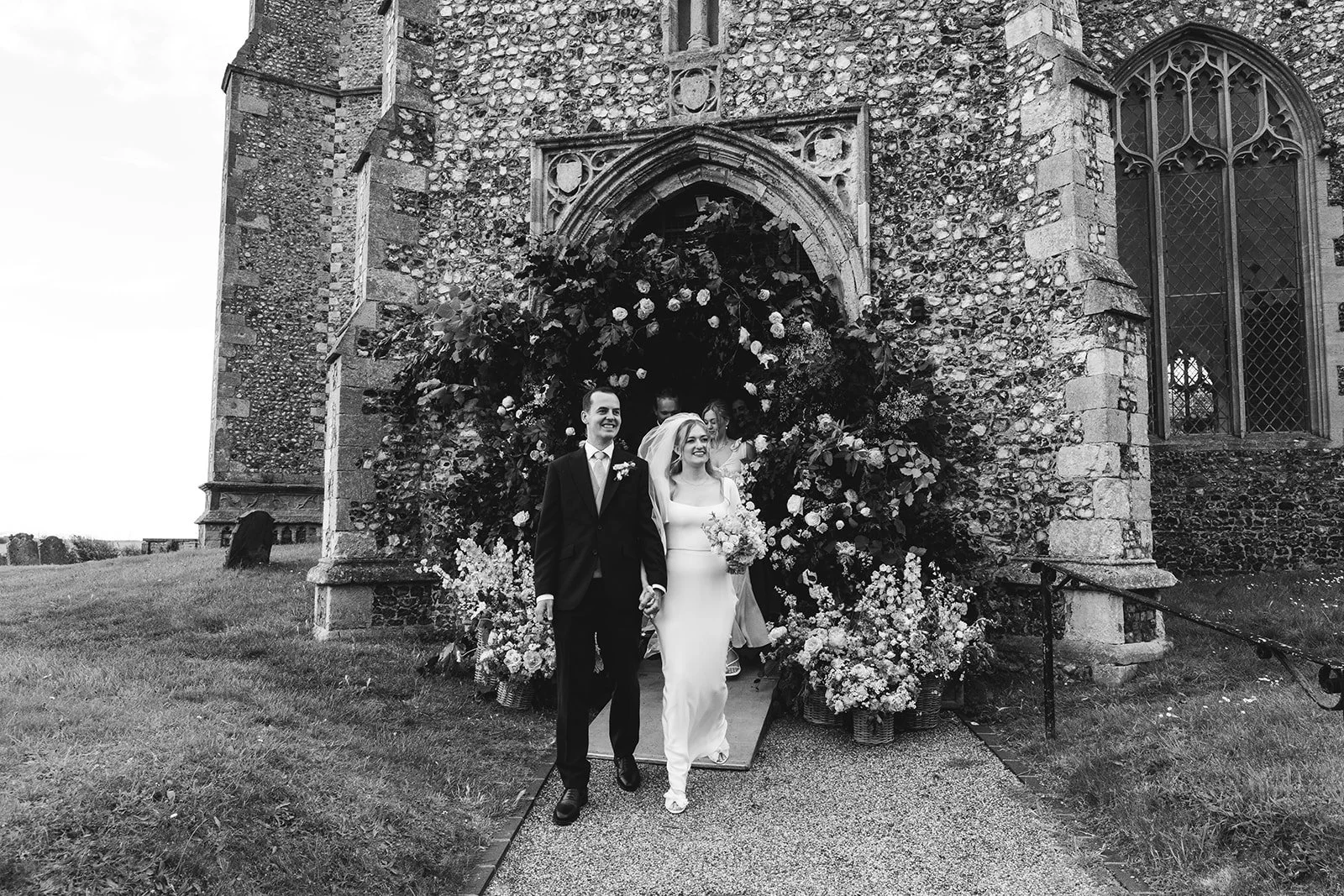 Black and white photo of a wedding couple exiting a church decorated with flowers. The bride is in a white gown holding a bouquet, and the groom is in a dark suit. They are holding hands and smiling.