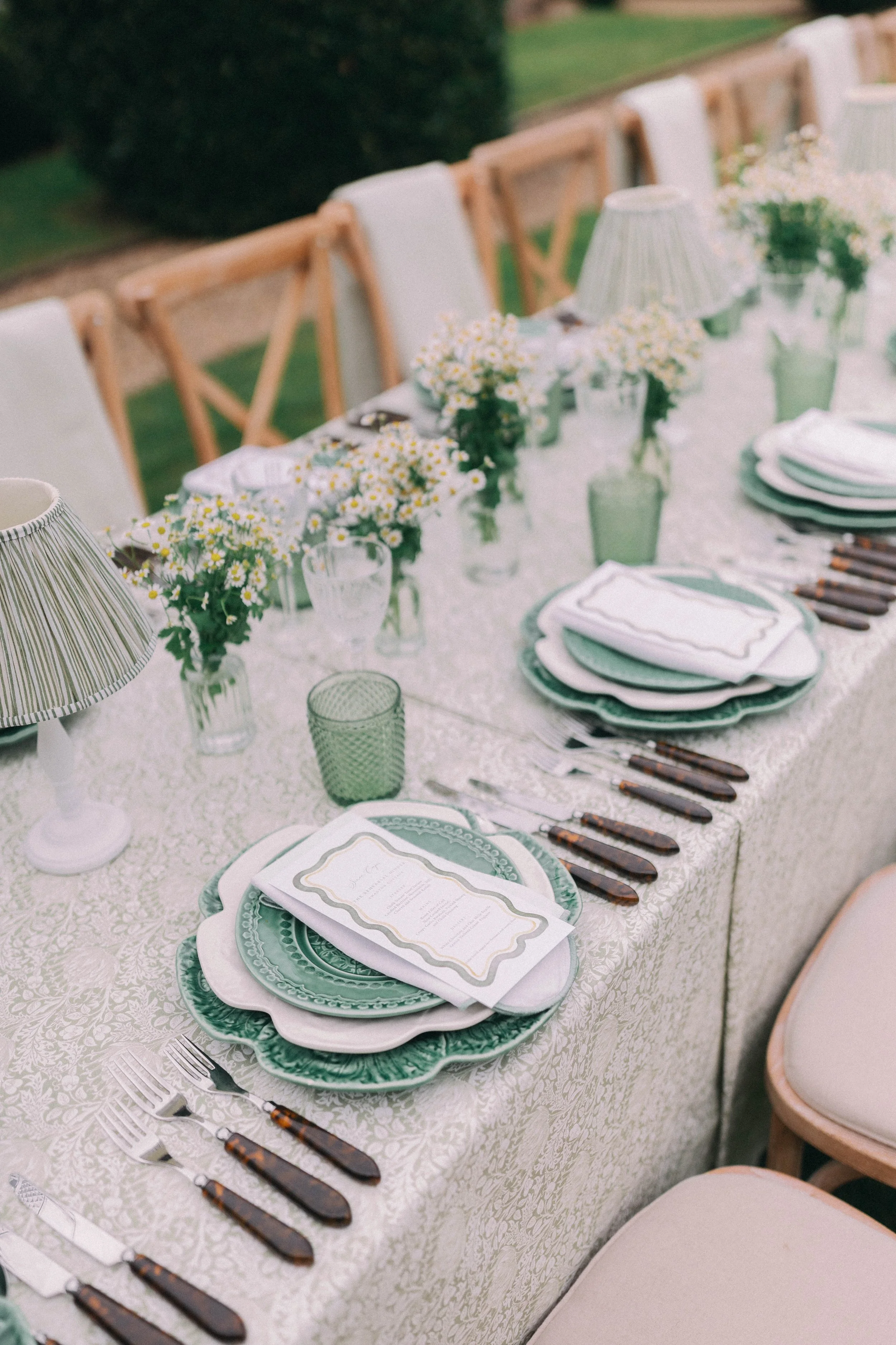 Elegant outdoor dining table set with green and white patterned plates, white napkins, vintage cutlery, floral centerpieces, and table lamps, with wooden chairs. Hosted in Norfolk planned by Attabara Studio