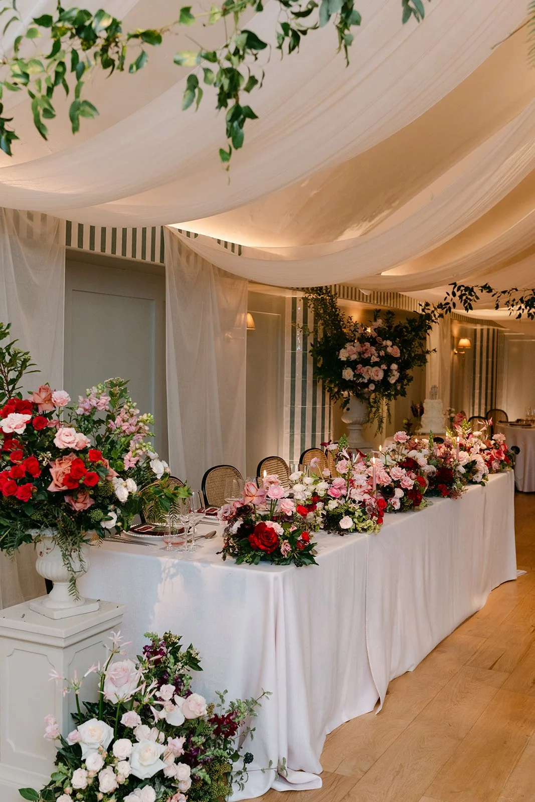 Elegant banquet table decorated with pink, white, and red flowers, set with glassware and silverware, in a room with draped ceiling fabric and soft lighting.