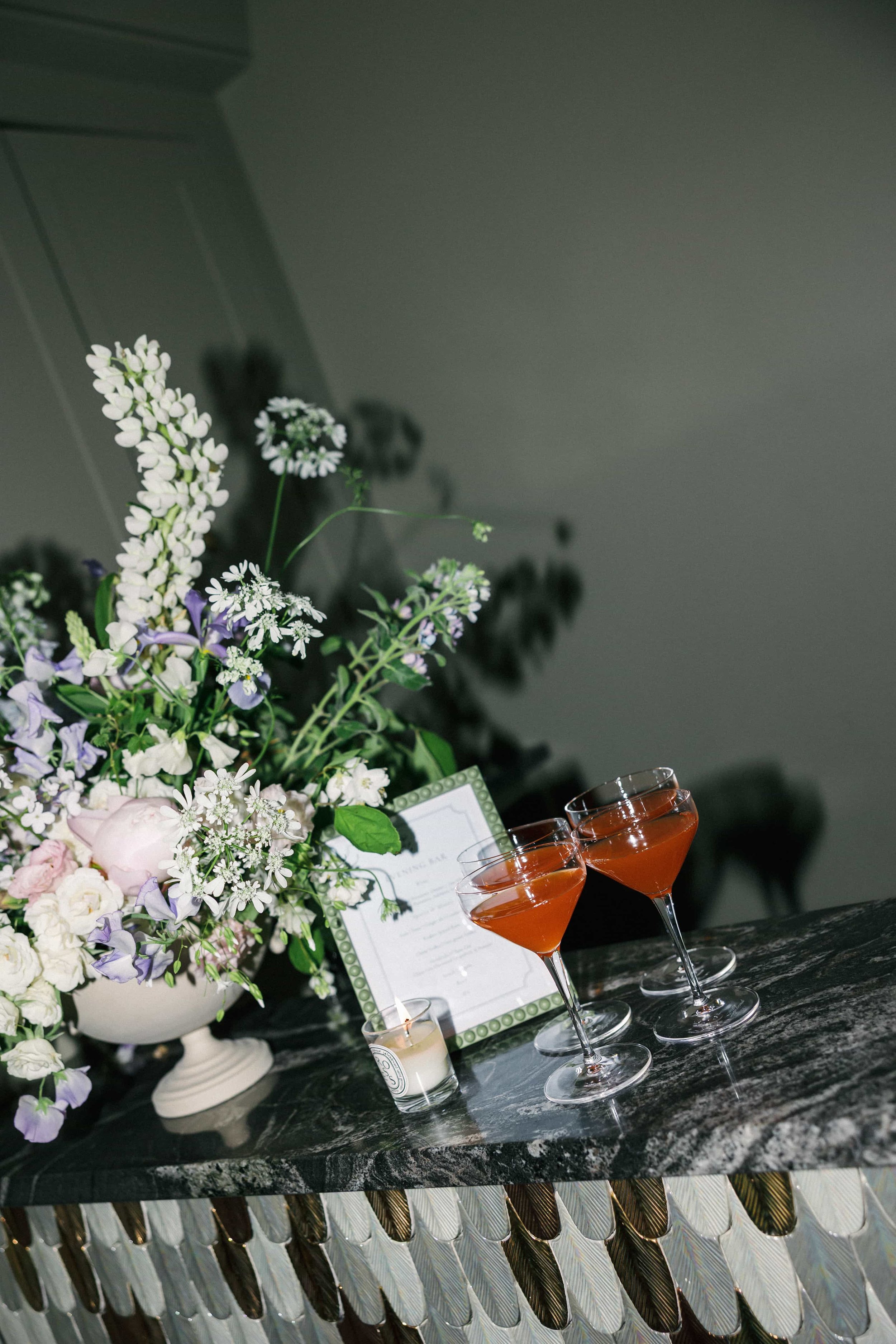 Elegant floral arrangement with white, purple, and pink flowers, two glasses of rose-colored cocktails, a lit candle, and framed menu on a dark marble tabletop.