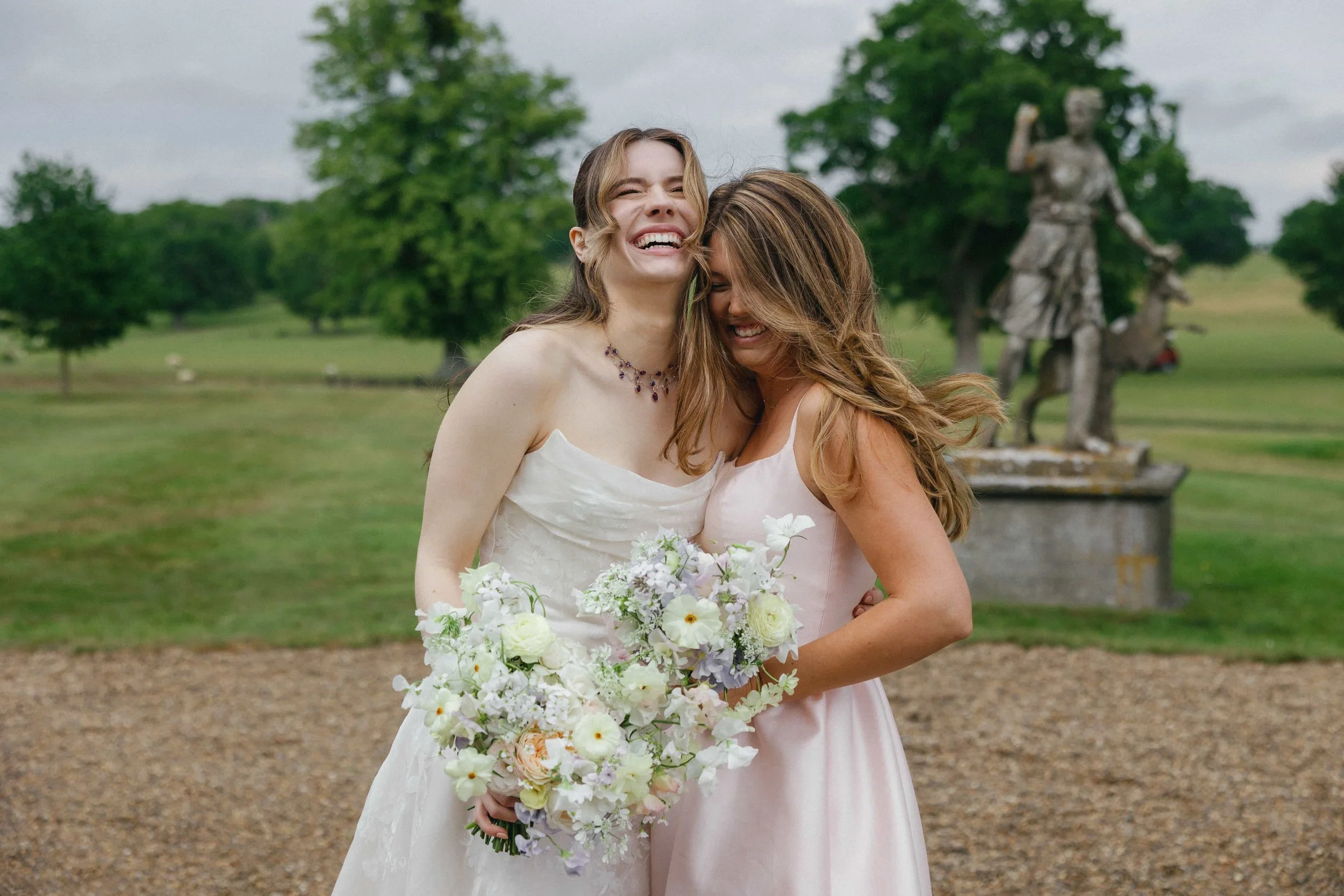 Two women in wedding attire sharing a joyful hug outdoors, with one holding a bouquet of flowers, and a statue of a woman on horseback in the background surrounded by greenery. The Botany House Florist at Wilderness Reserve, Planned by Attabara