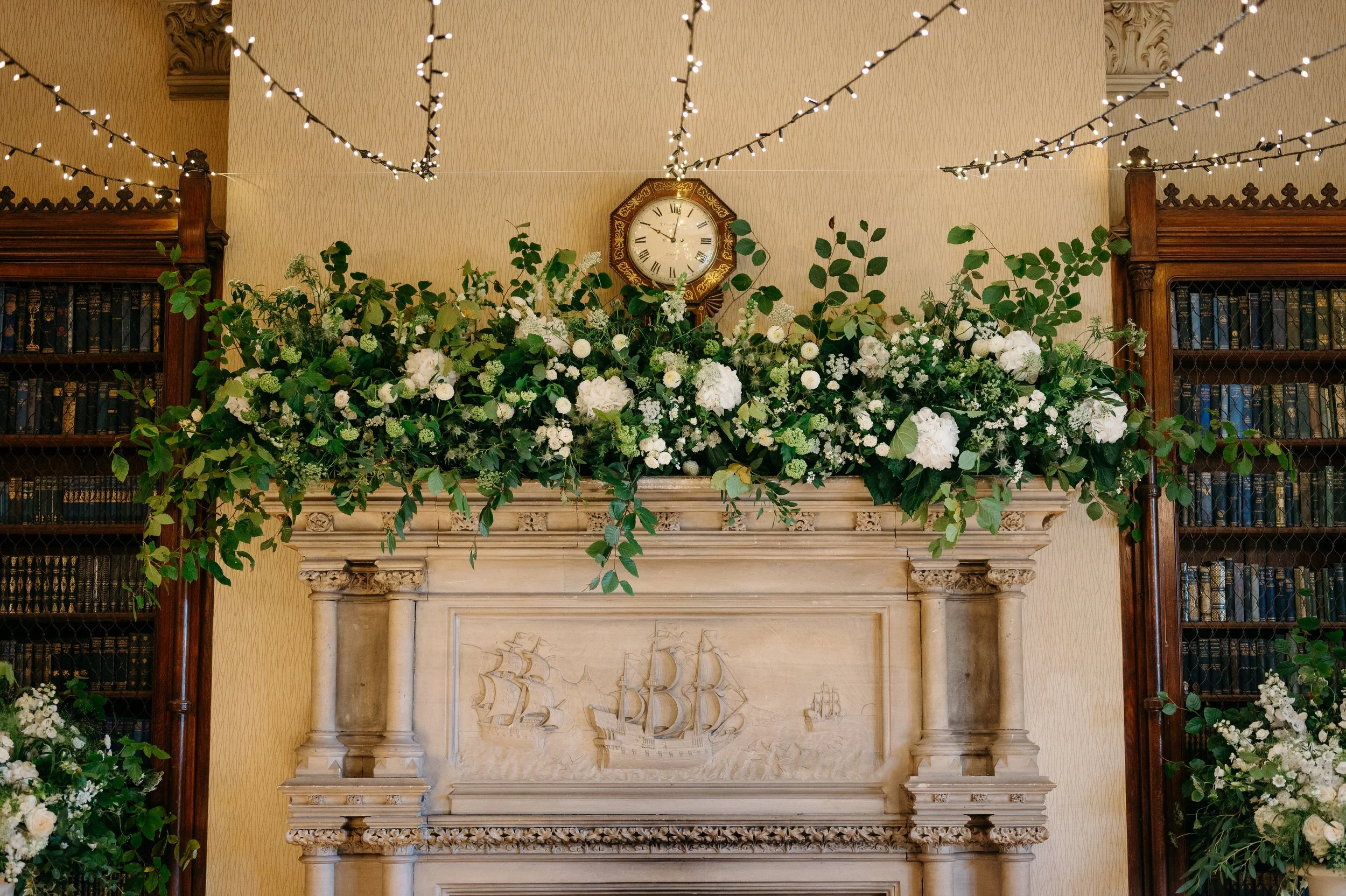 Decorative fireplace mantel adorned with white flowers and greenery, with a clock, bookshelves on either side, and string lights overhead.