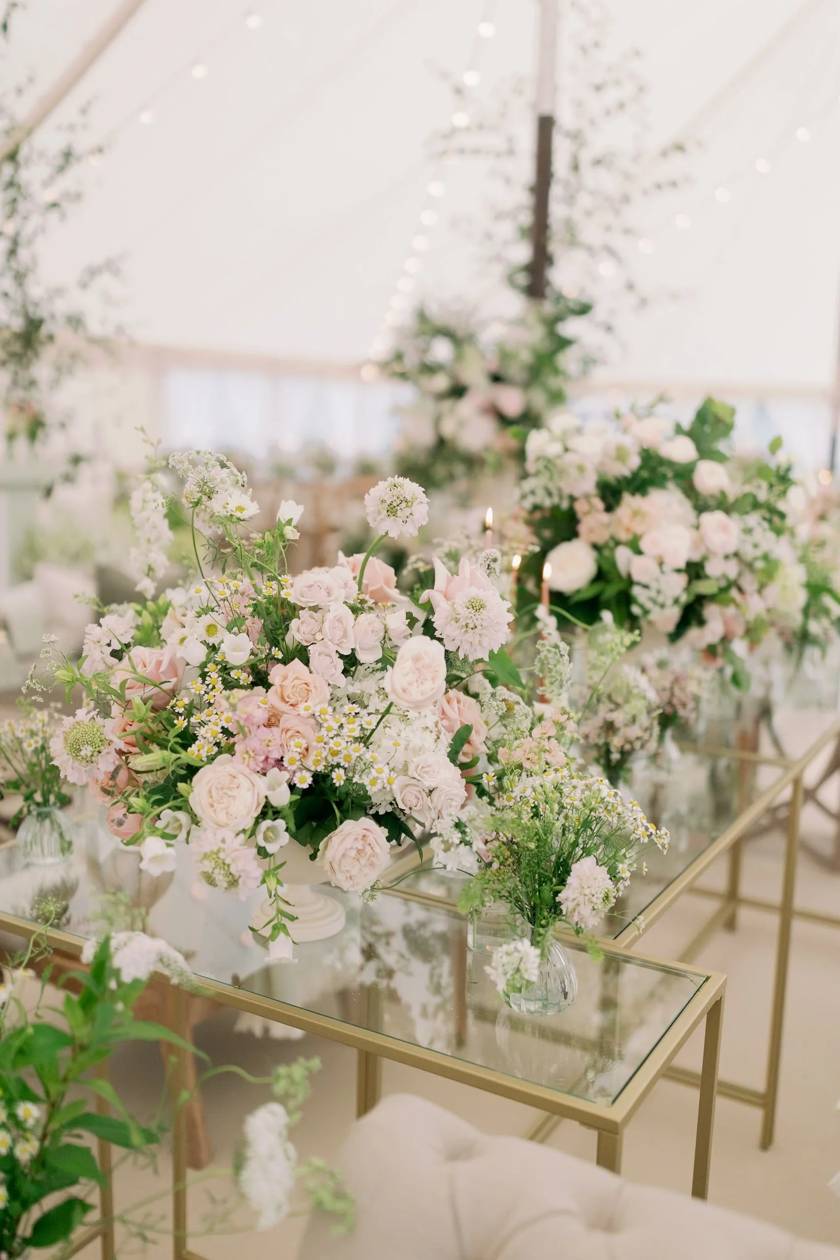 Elegant wedding centerpiece with soft pink and white flowers arranged on a glass table, surrounded by additional floral arrangements in clear vases, in a bright, sail and peg sailcloth marquee