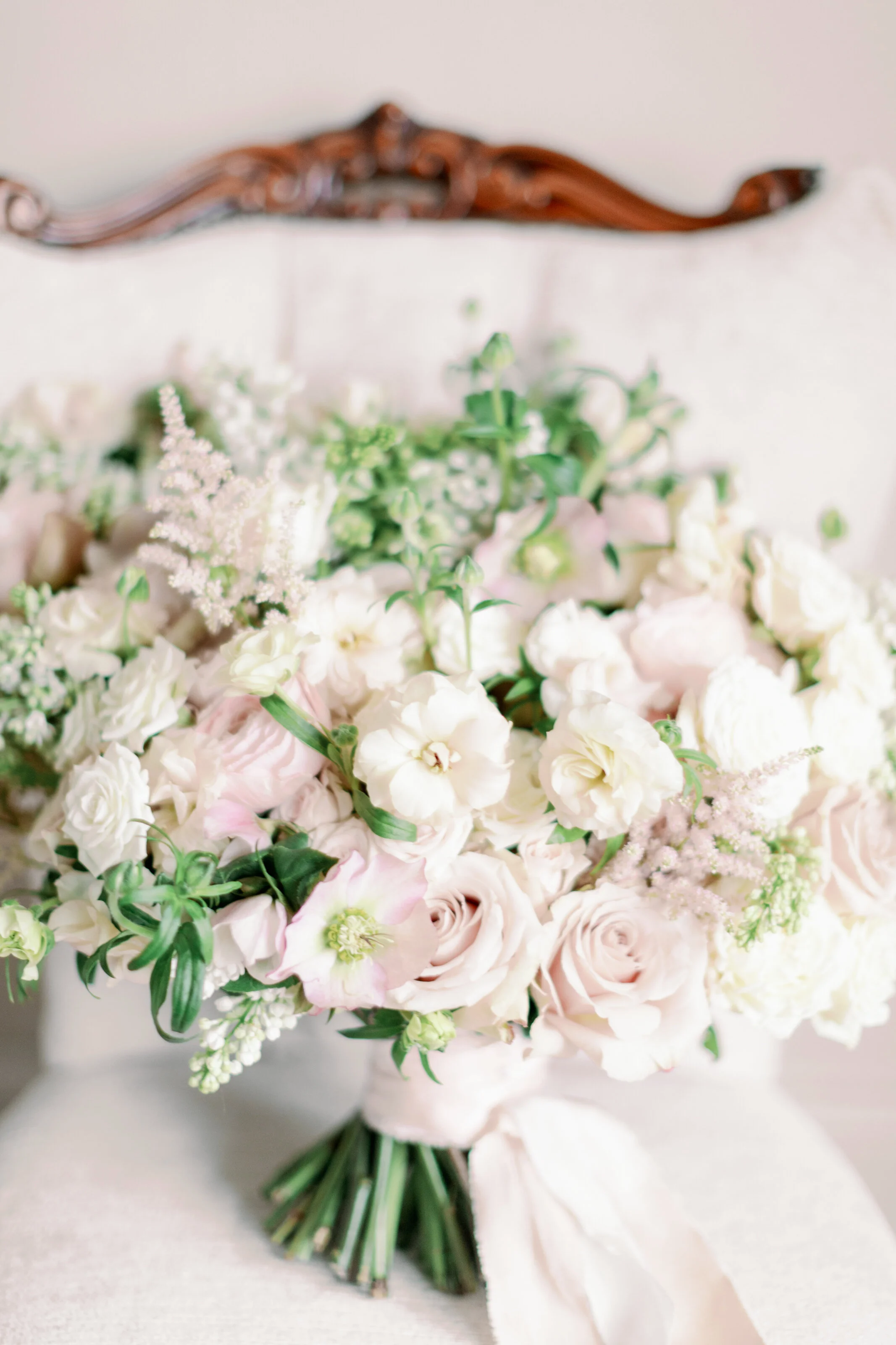 Elegant bouquet of white and pale pink roses, lisianthus, and greenery resting against a cream-colored upholstered chair.